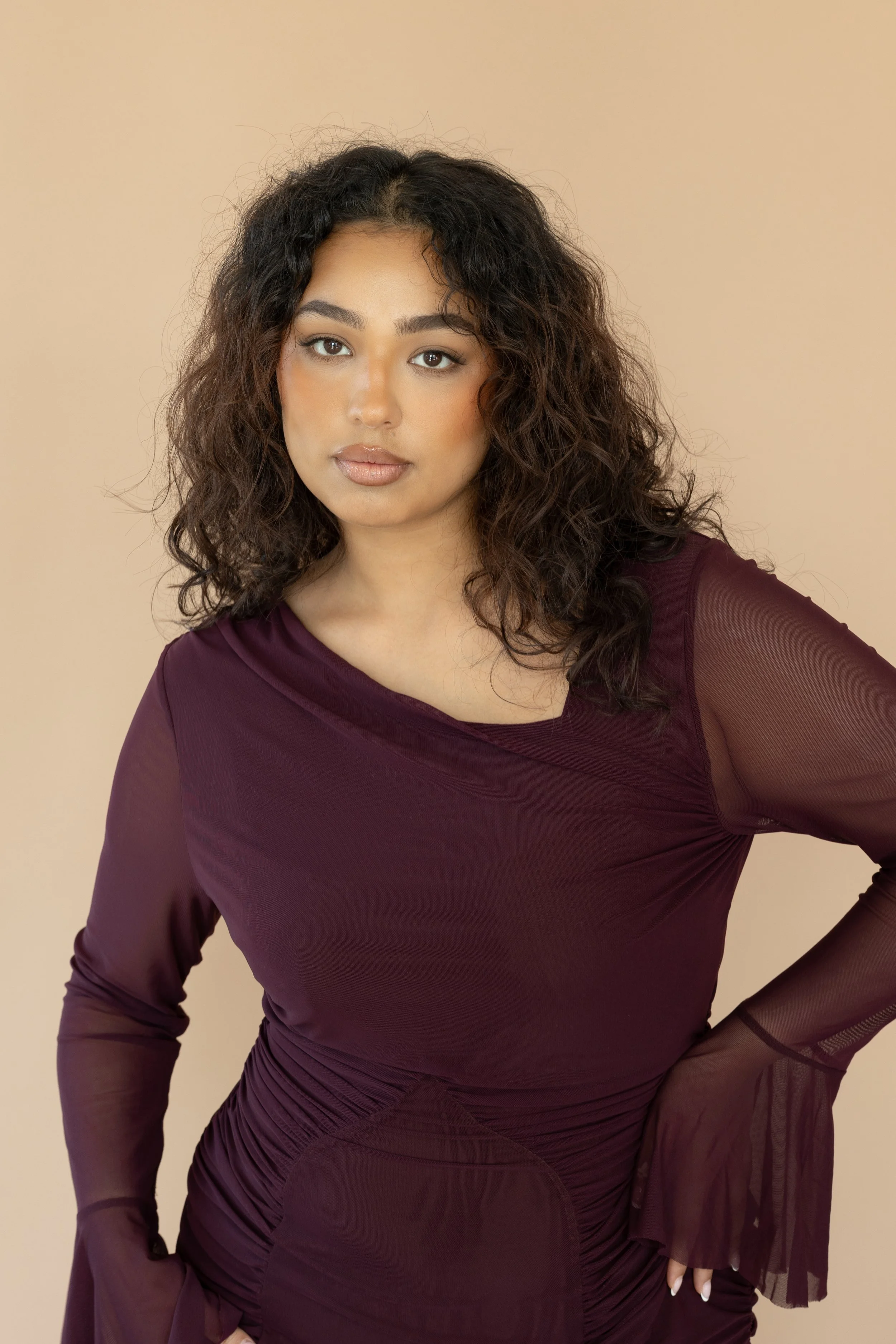 A woman with wavy dark hair wearing a long-sleeve, dark purple dress with sheer sleeves and gathered details at the waist, standing against a beige background.