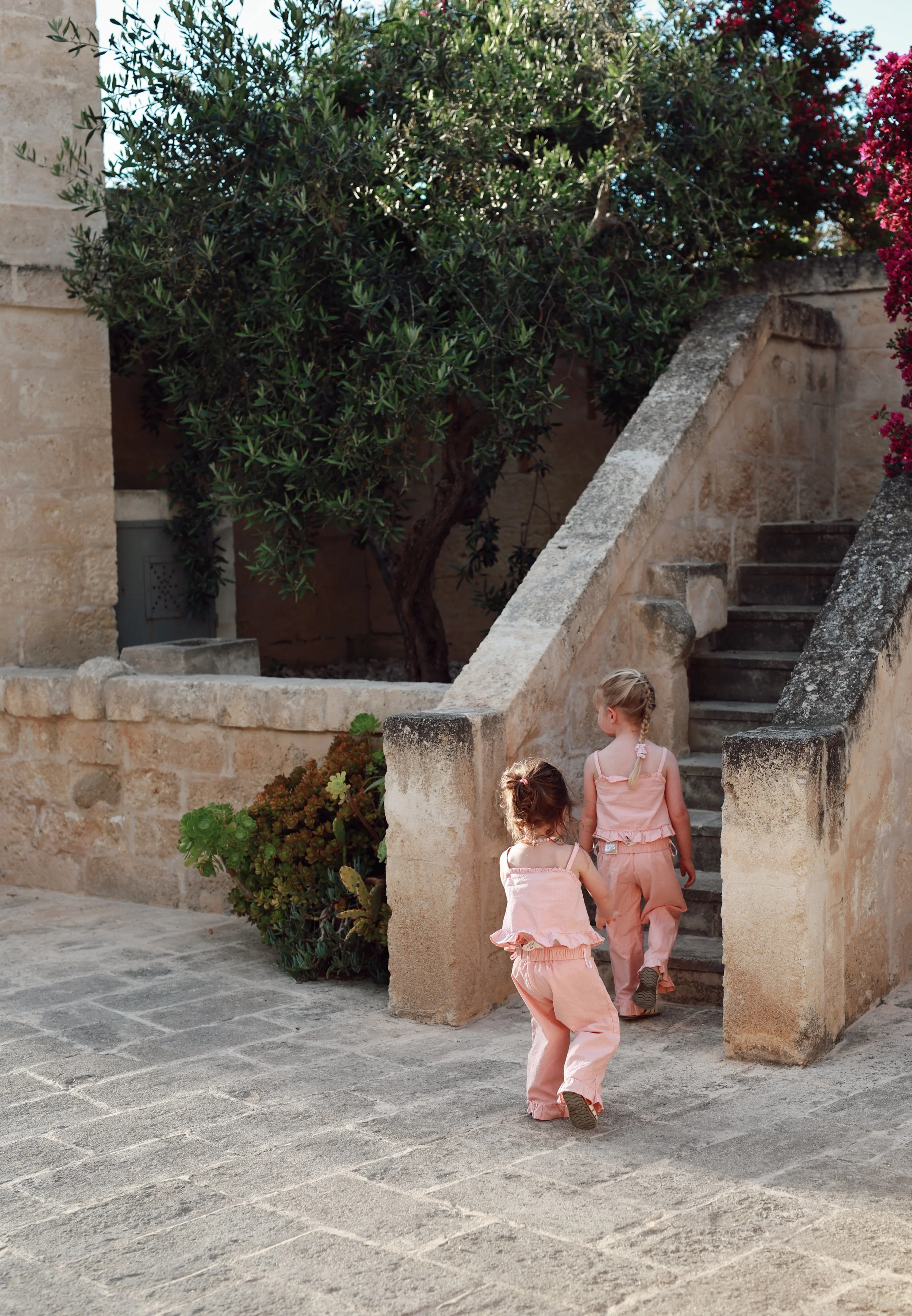 Three children in pink outfits walking up stone stairs outside, near a stone wall with plants and trees.