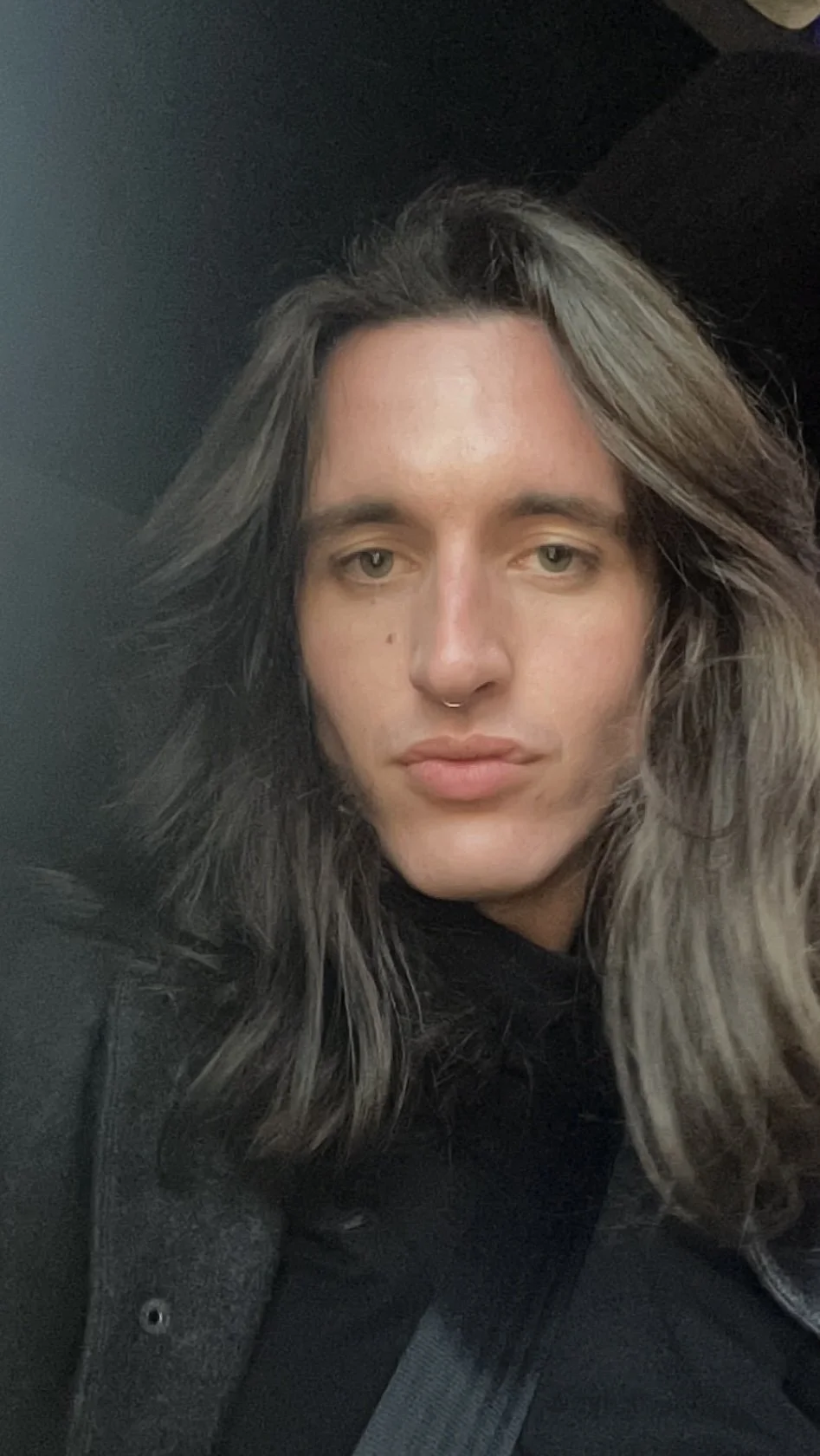 A person with light skin and long, wavy, grayish-brown hair taking a selfie indoors.