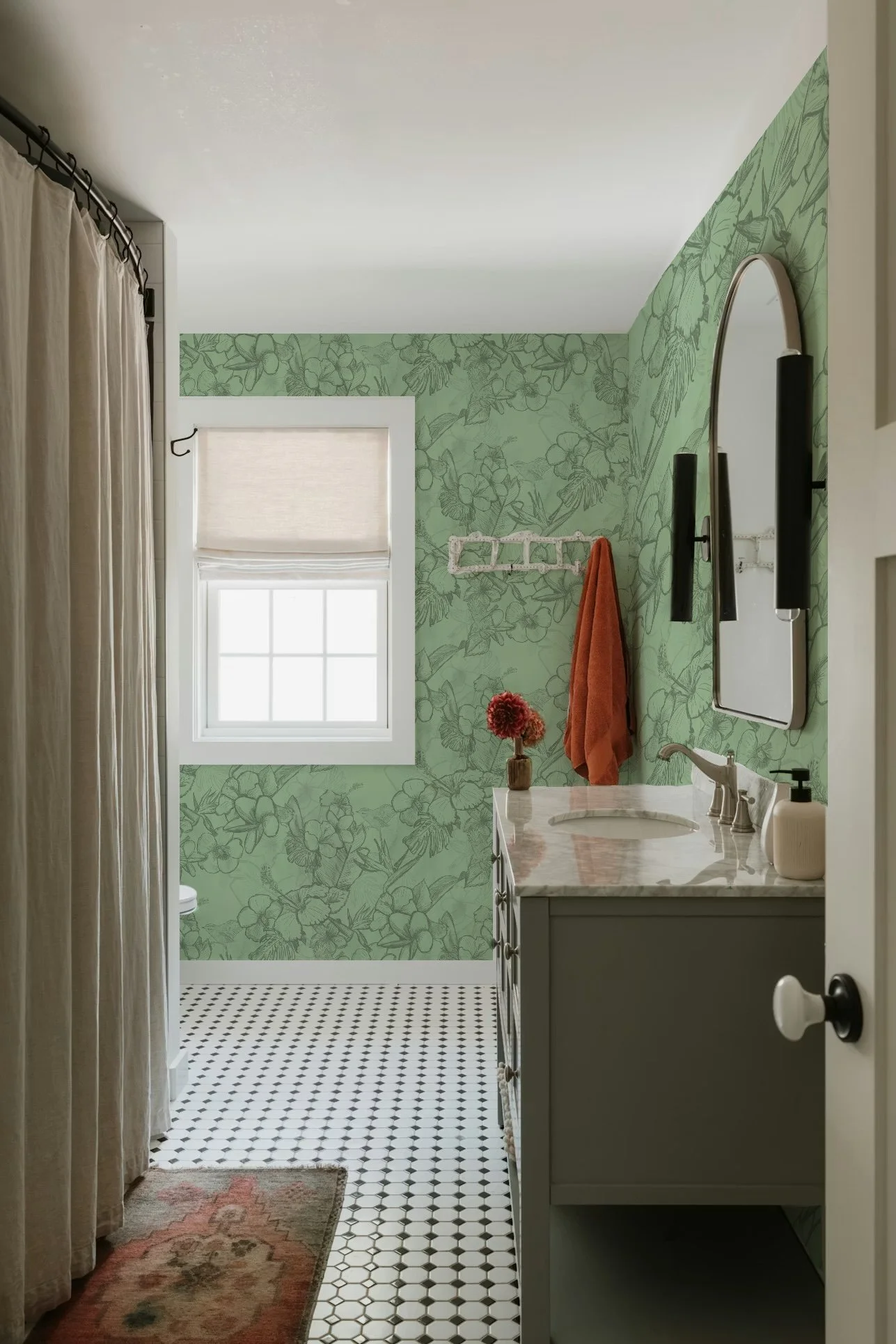 A bathroom with green floral wallpaper, a window with a white Roman shade, a marble countertop vanity with a sink, a round mirror, and orange towels hanging on the wall.