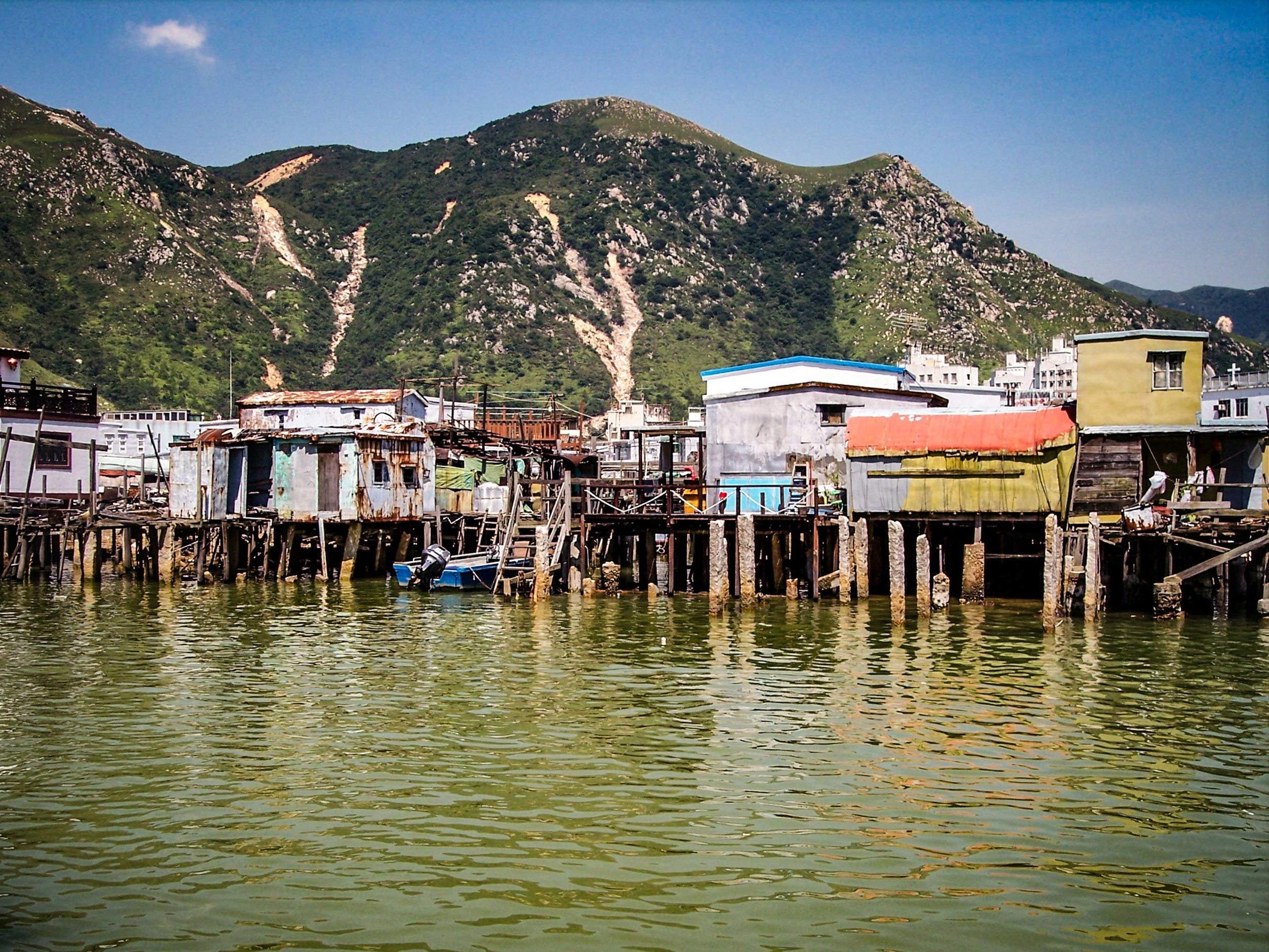 A view of a hillside with green vegetation and rocky patches behind a small fishing town with stilt houses along a waterway.