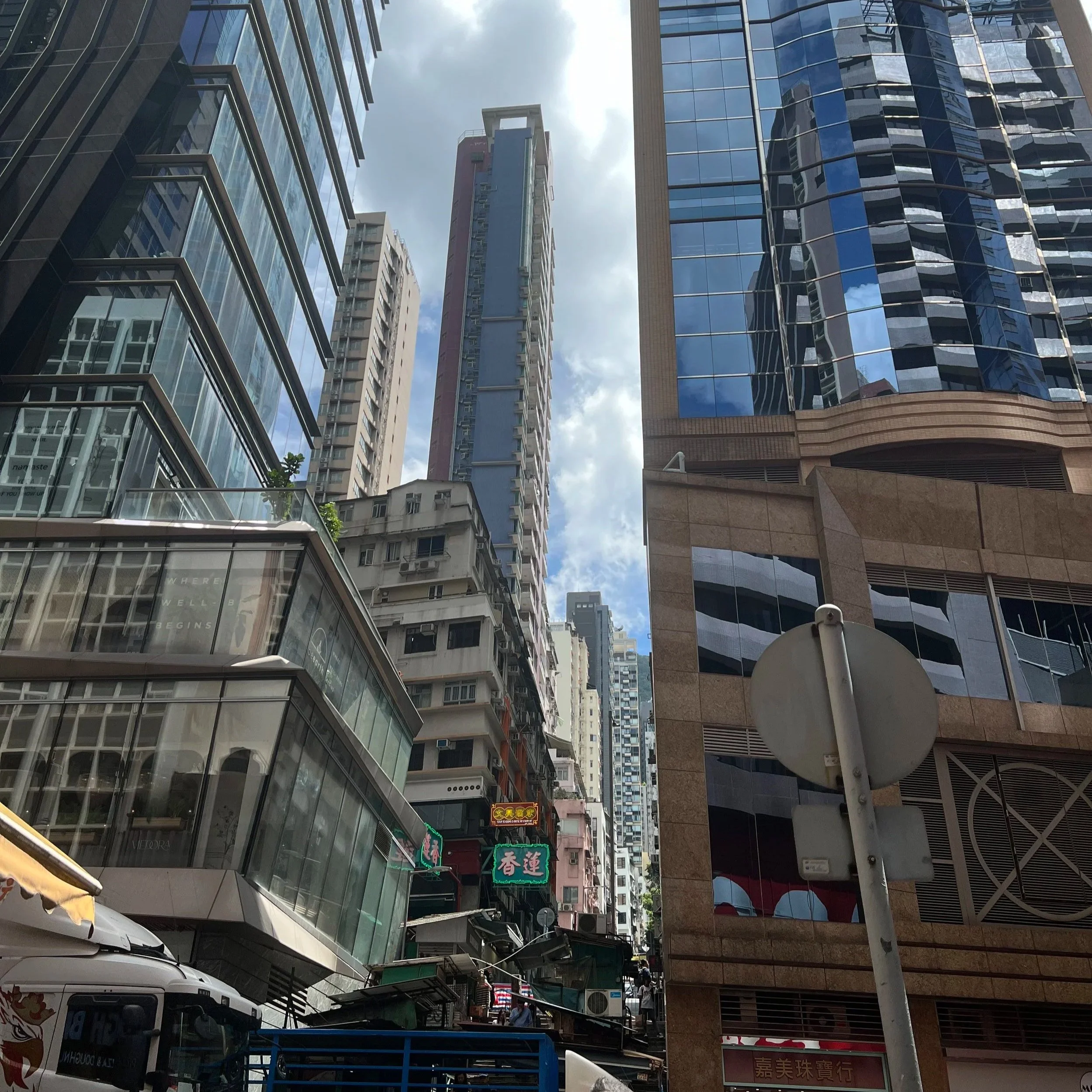 City street with tall modern glass buildings, signs in Chinese, parked cars, and street vendors, under partly cloudy sky.