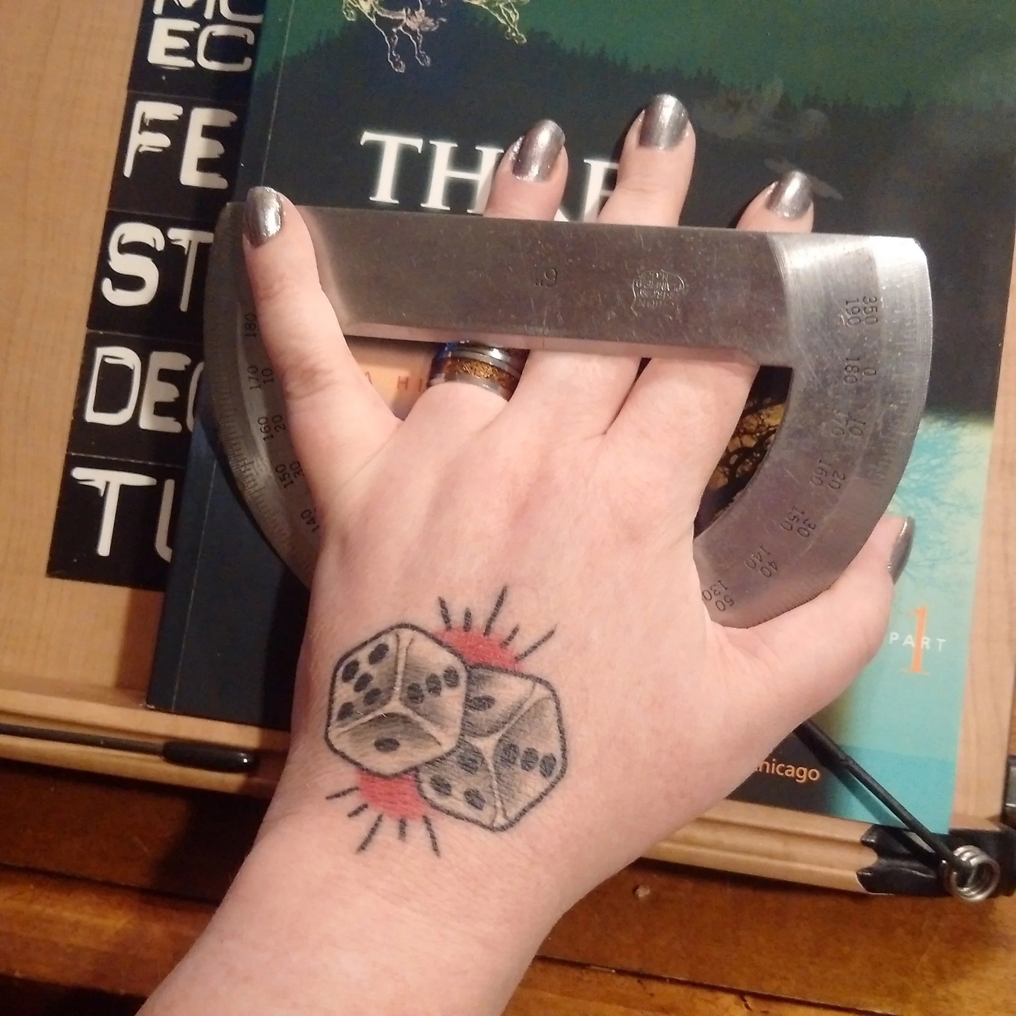 Marie's left hand, nails polished gunmetal grey, and a tattoo of dice showing a hard 8 on the back, holding a vintage metal protractor against a book on a wooden stand.