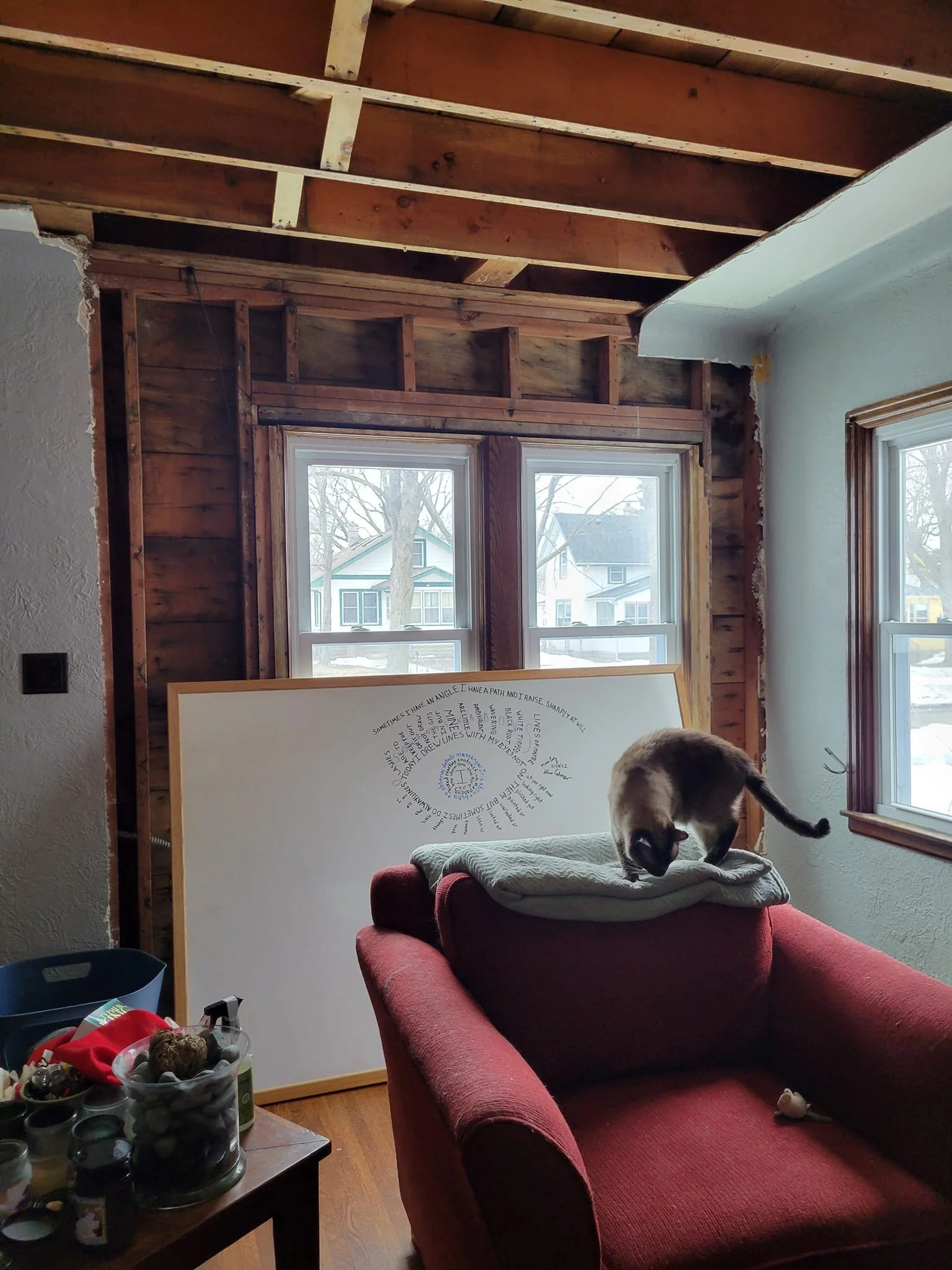 A living room after damage from an ice dam. A large dry erase board shows the draft of a concrete poem in the shape of an eye, a table holds miscellaneous displaced items, and a cat inspects a burgundy chair. Wall & window exposed to the studs.