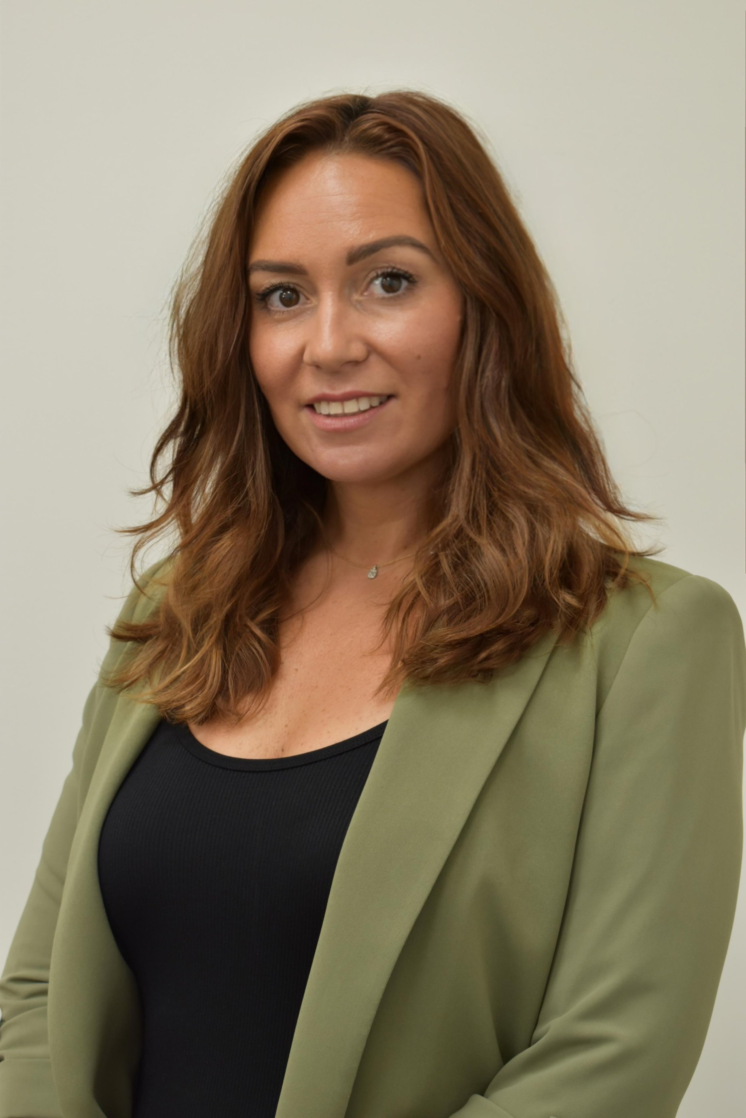 A woman with long, curly auburn hair and brown eyes smiling, wearing a black top and an olive green blazer, standing in front of a plain, off-white background.
