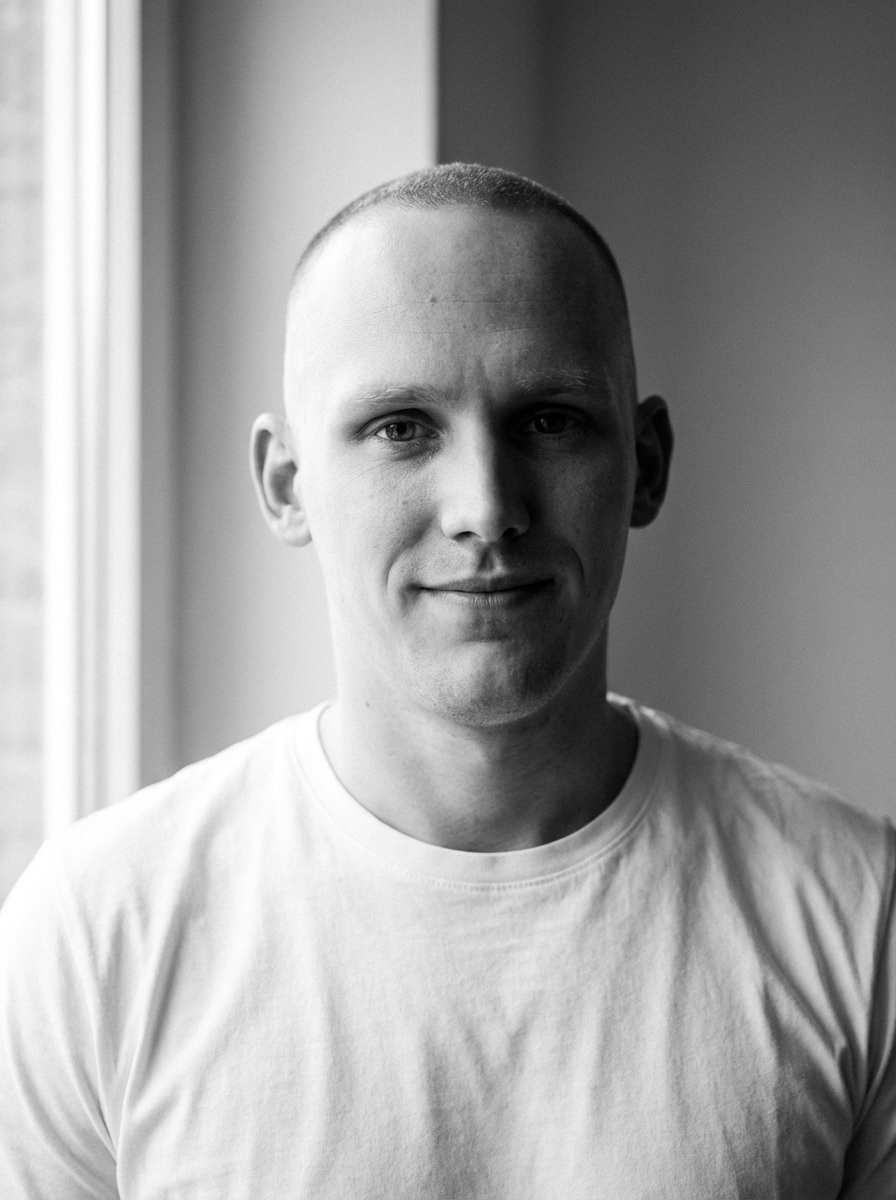 Black and white close-up portrait of a young man with a buzz cut, standing indoors near a window, wearing a plain white t-shirt, smiling subtly.