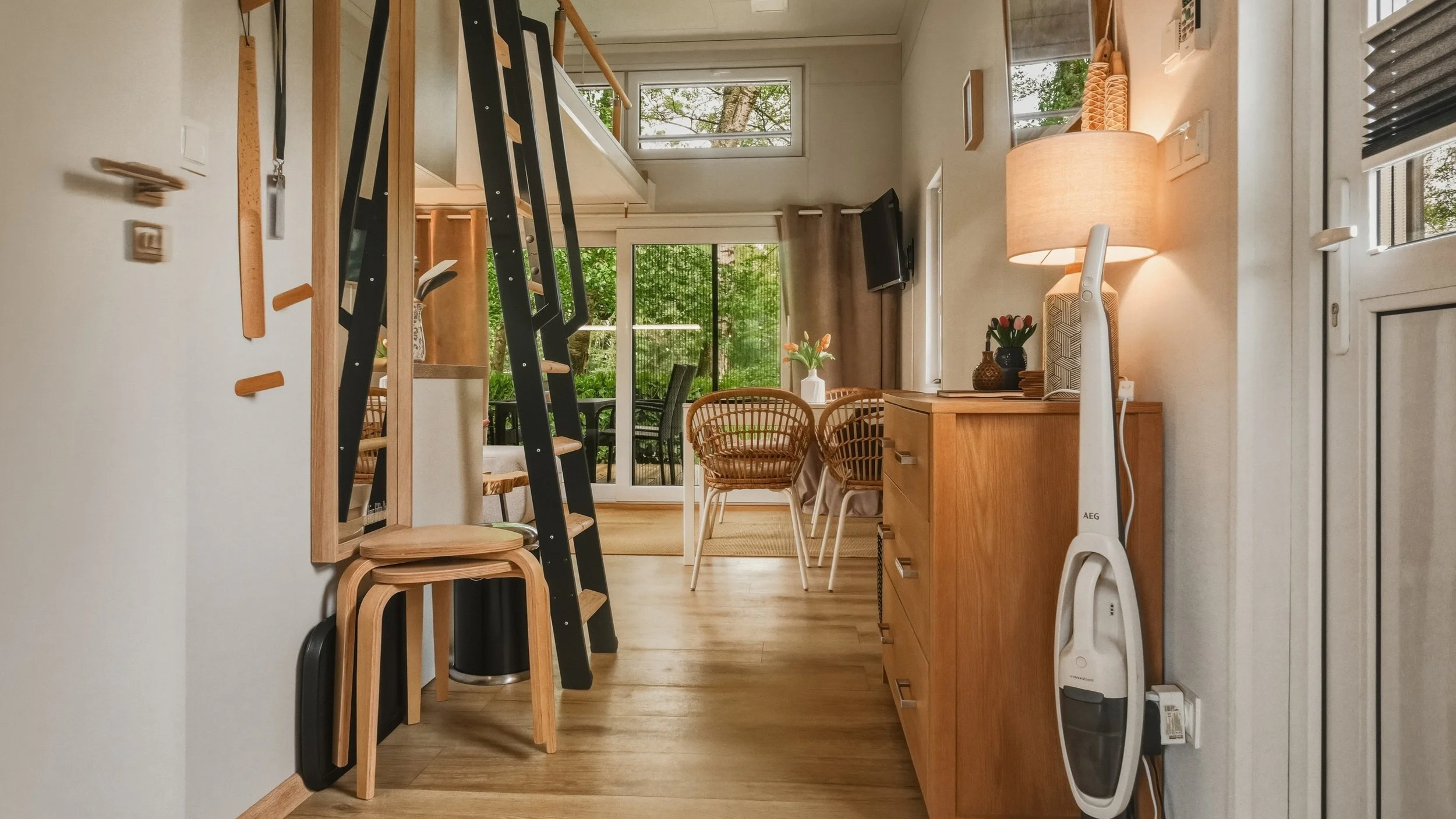Living room with large windows and sliding glass door, wooden dining table with chairs, a TV mounted on wall, a sideboard with a table lamp, a vacuum cleaner, and outdoor patio furniture visible outside.