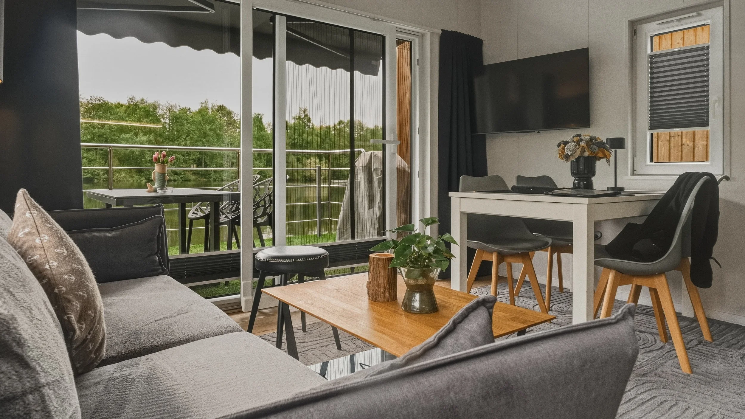 Living room with sliding glass door leading to balcony with trees, a TV on wall, a dining table with chairs, and a couch with pillows.