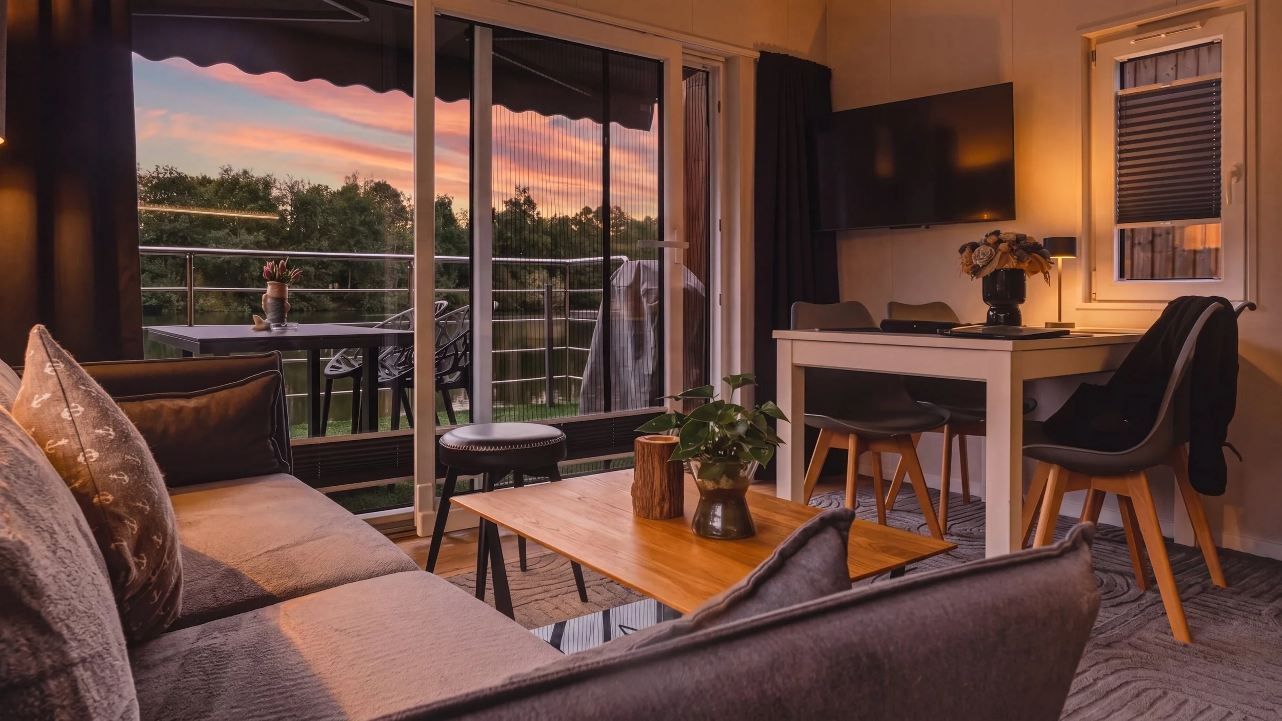 Living room with large sliding glass doors leading to a balcony, showing a sunset sky with pink and purple clouds over a landscape of trees.