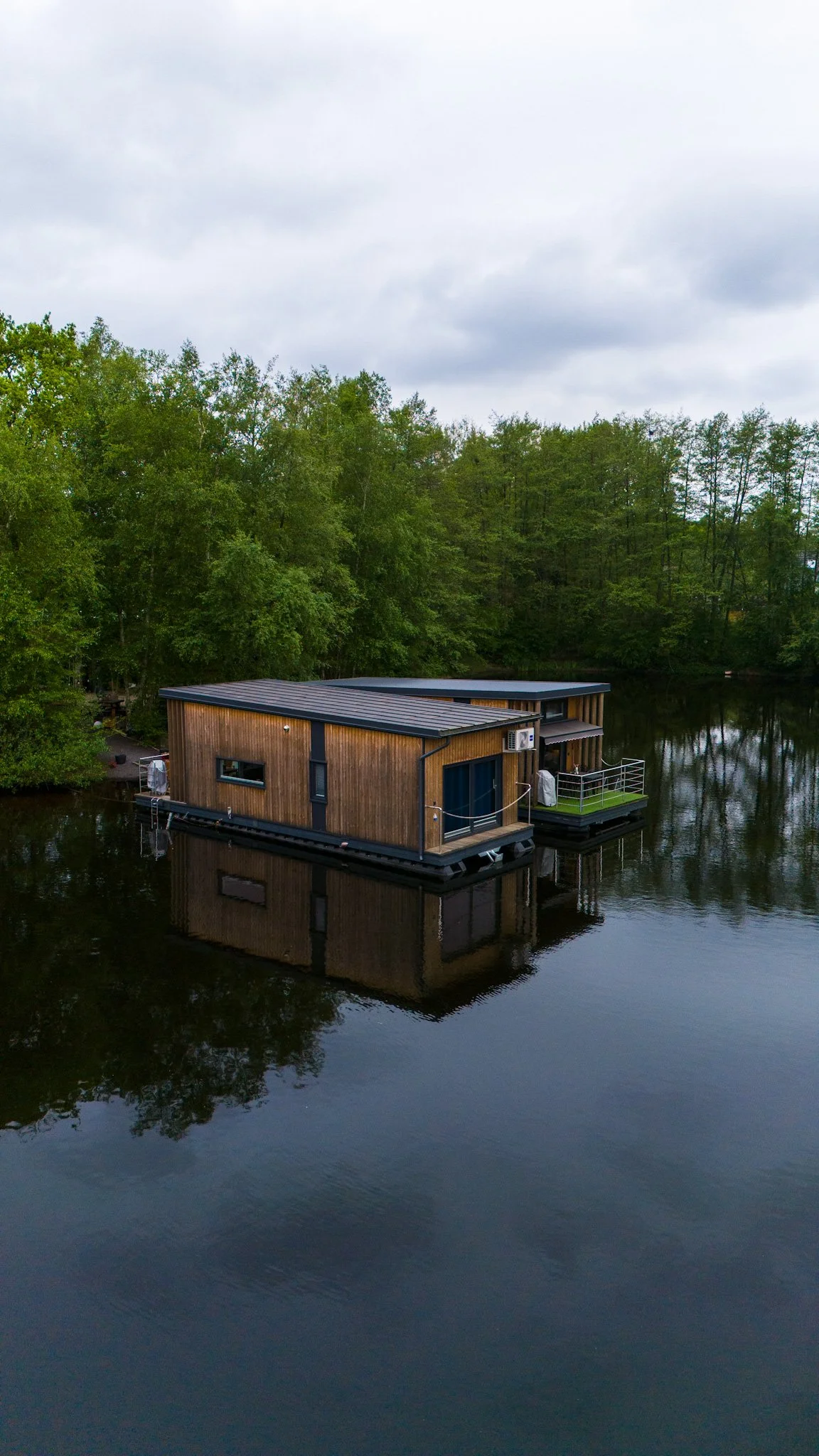 A modern wooden houseboat floating on a calm river surrounded by dense green trees under an overcast sky.