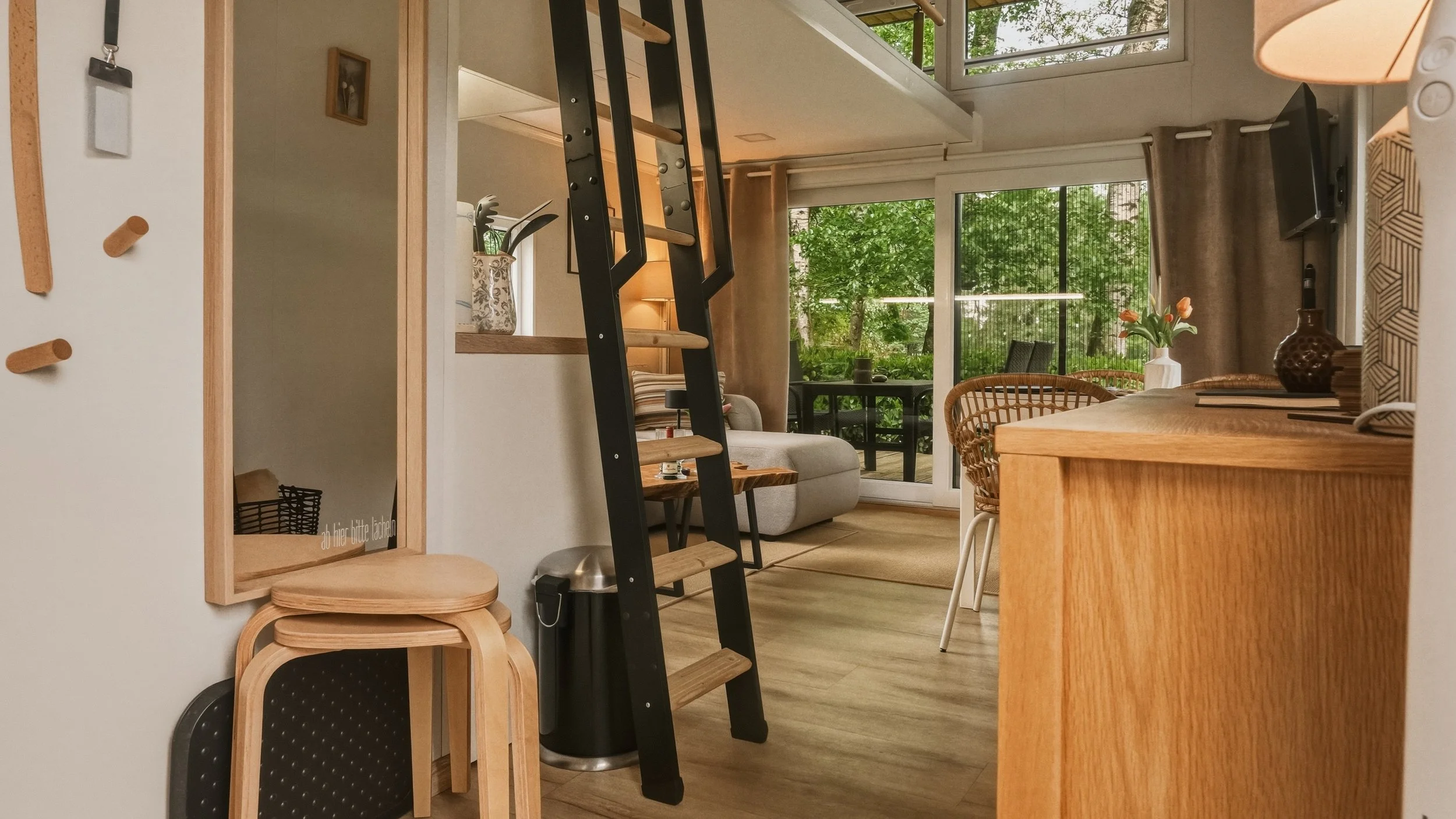 A cozy living room with a view of lush greenery outside. The space features a small table with a vase of flowers, a sofa, a dining table with chairs, large windows, and a staircase with black and wooden steps leading up. There is also a mirror on the wall and a lamp.