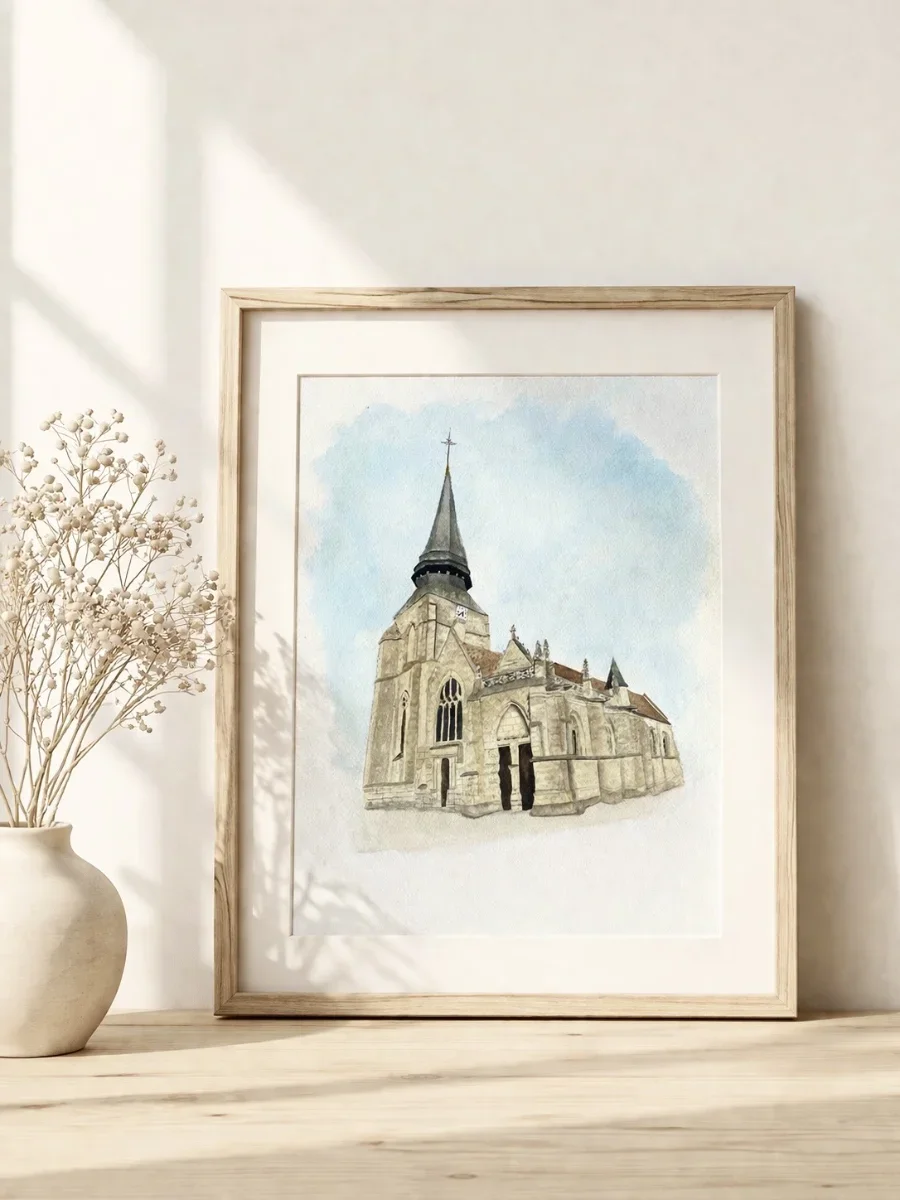 Une peinture aquarelle représentant une église avec une tour pointue, dans un cadre en bois clair, posée sur une table en bois clair, entourée de quelques objets décoratifs blancs, dont une vase avec des fleurs séchées et des bougies.