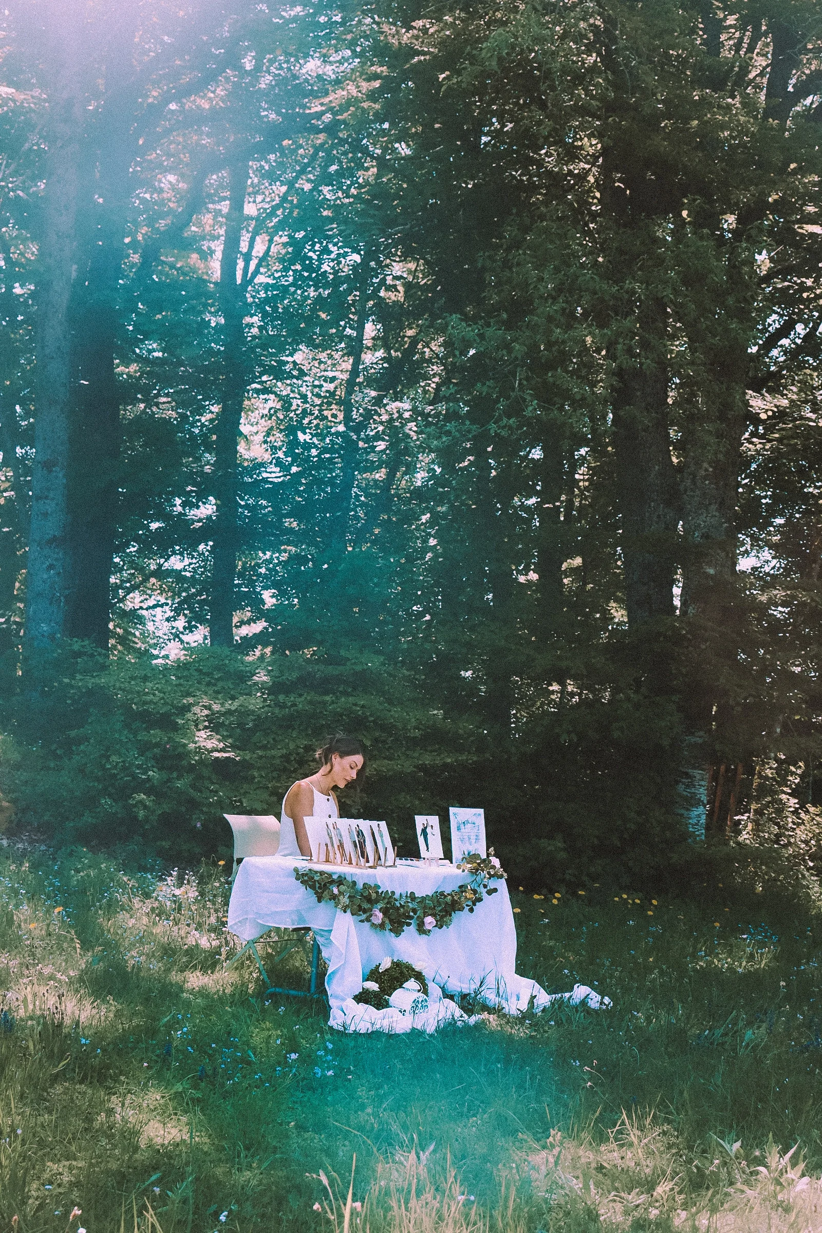 Une femme se trouve à une table blanche décorée de fleurs, dans un environnement boisé en plein air, probablement lors d'un événement ou marché artisanal, avec des dessins ou œuvres d'art exposés sur la table.