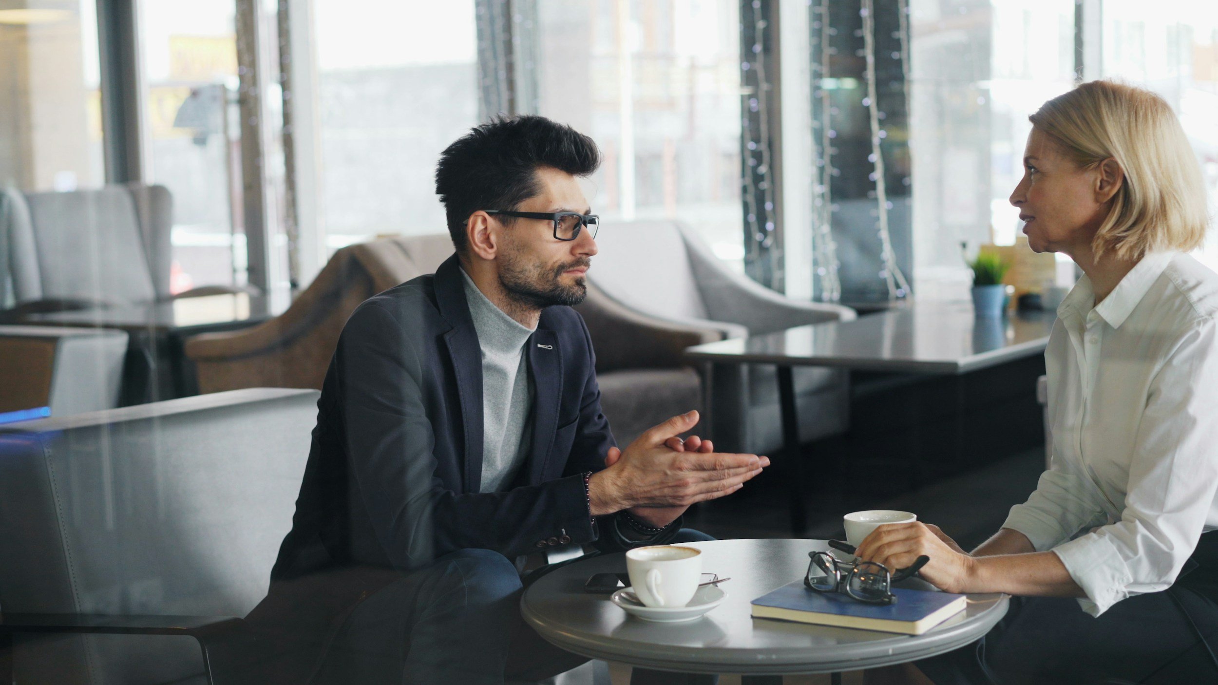 A man and woman having a serious conversation at a coffee shop, with cups and books on the table.