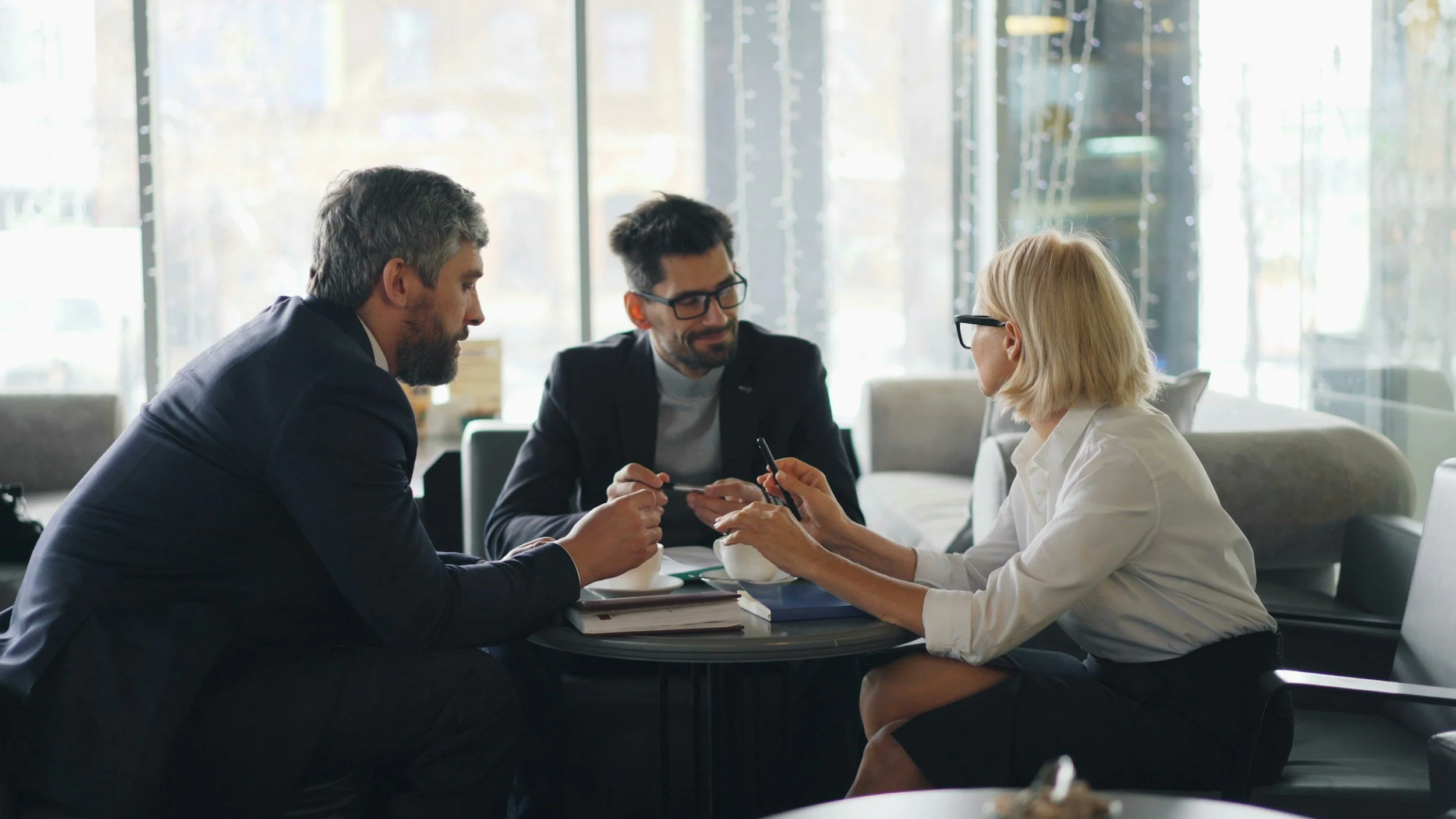 Three business professionals, two men and one woman, engaged in a discussion at a table in a modern office lobby with large windows and natural light.