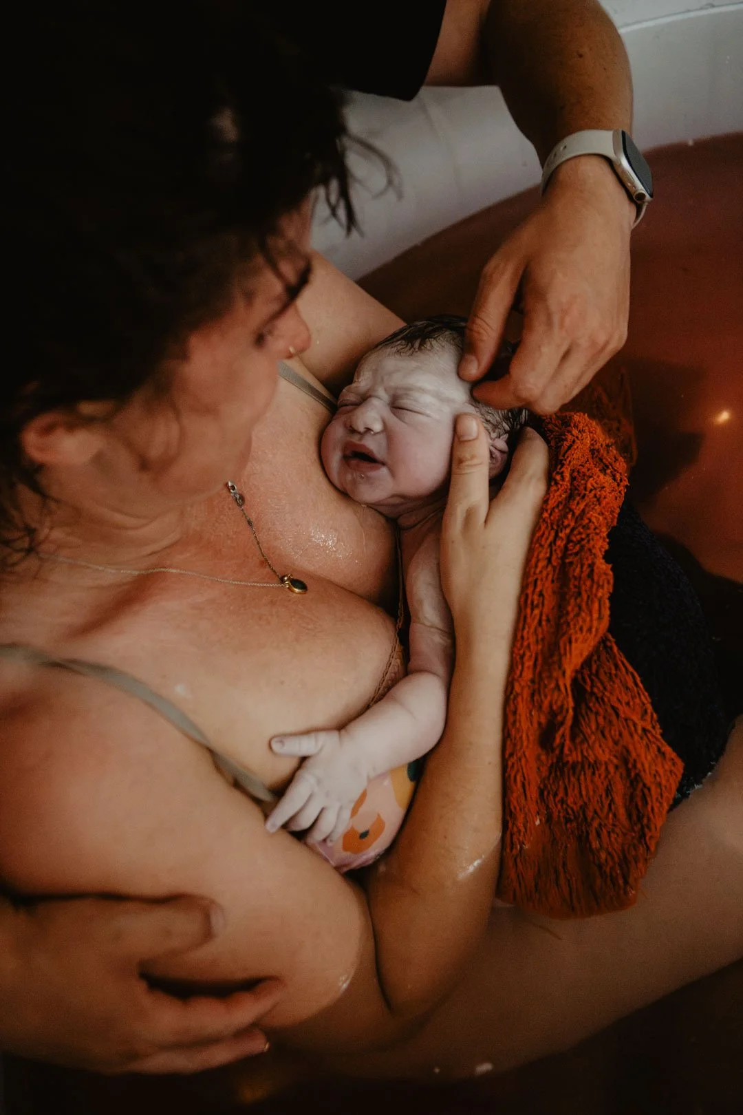 A woman is giving birth in water, holding a newborn baby close to her chest after delivery, with a person helping in the process.