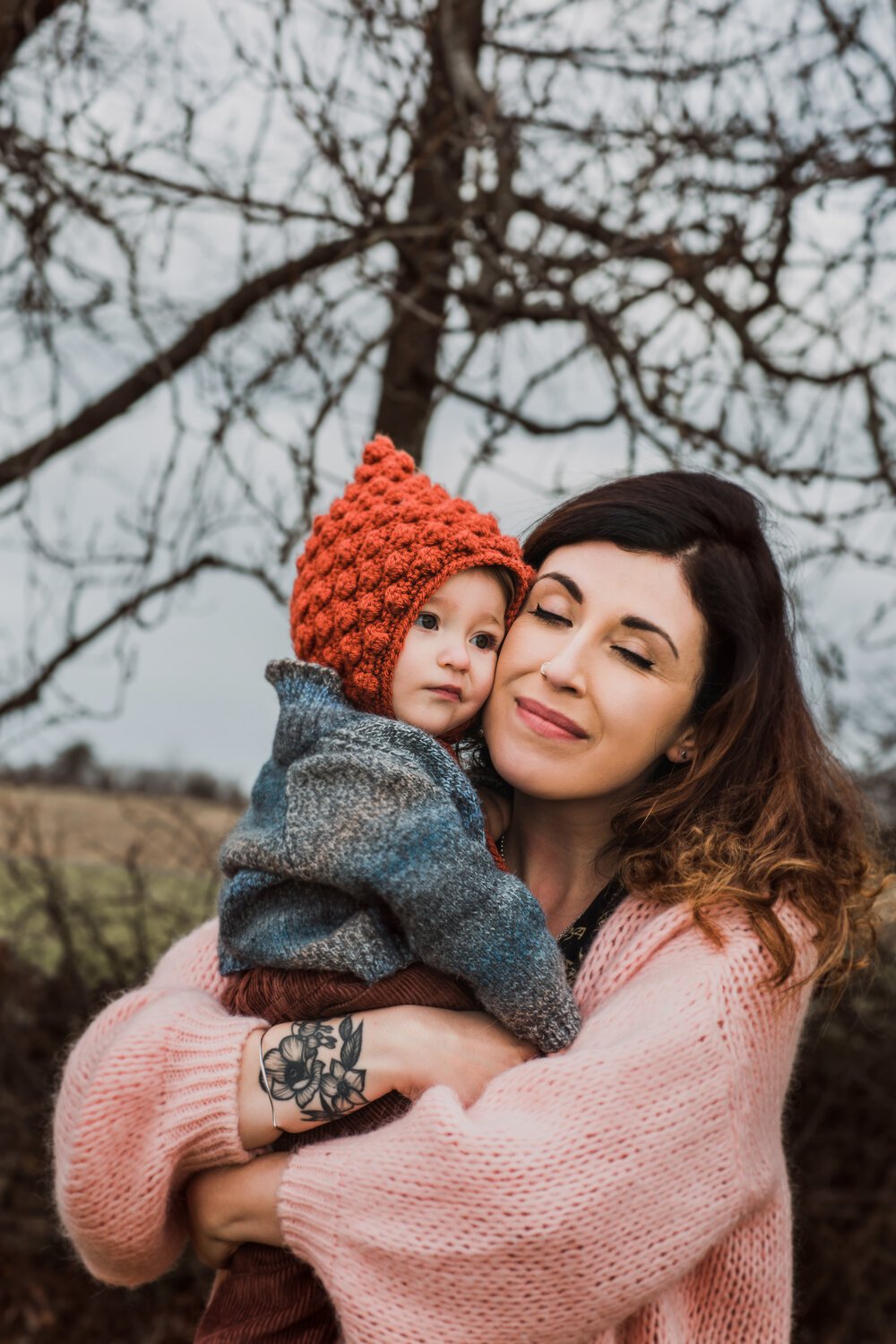 Woman hugging a young child outdoors during winter; woman has dark hair, is wearing a pink sweater, and has a tattoo on her wrist. The child is wearing a red knitted hat and a gray sweater, and looks slightly away. Bare trees and a cloudy sky are in the background.
