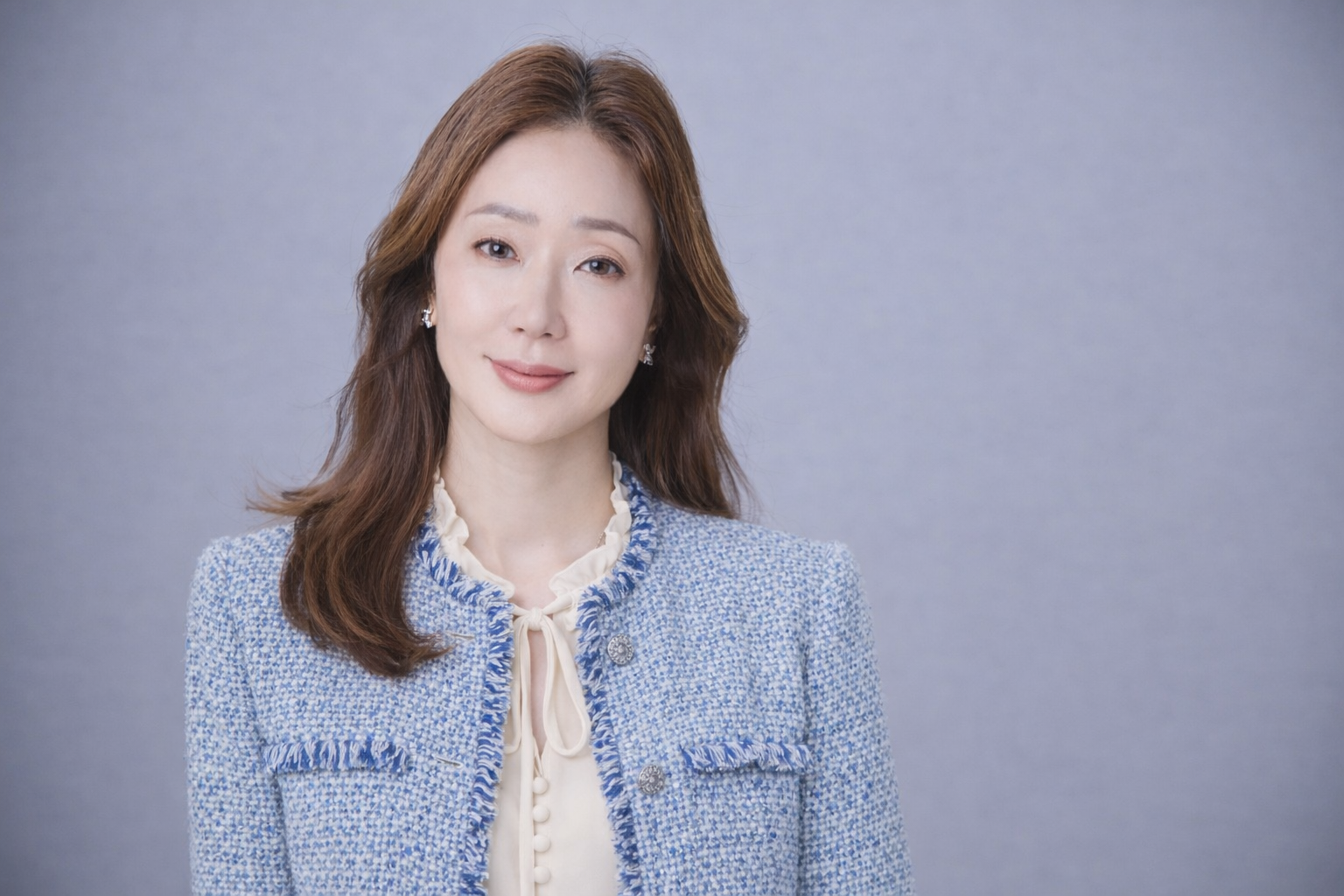 Portrait of a woman with shoulder-length brown hair wearing a blue tweed jacket and a cream blouse, standing against a plain gray background.