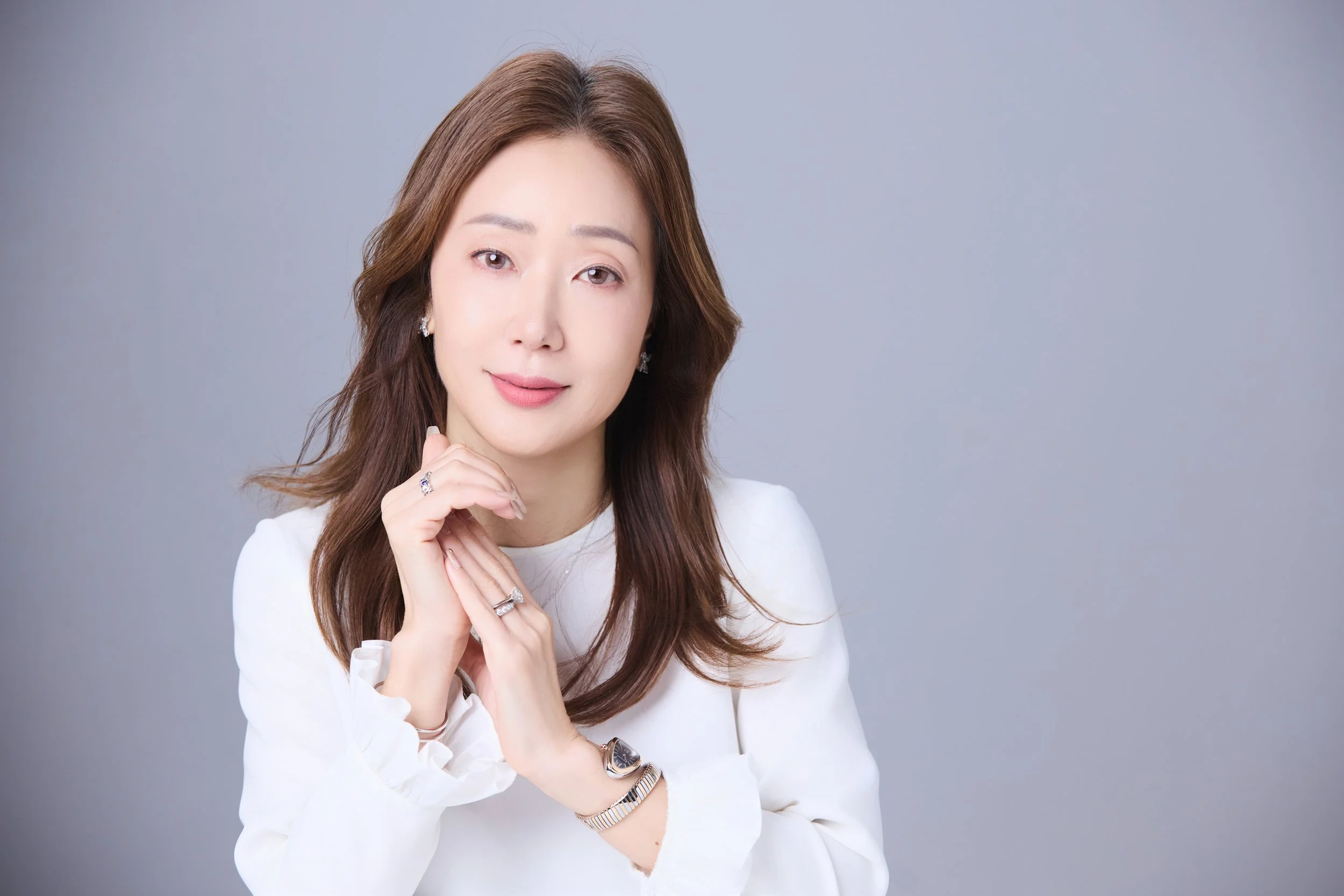 A woman with brown hair wearing a white outfit and jewelry, posing against a gray background.