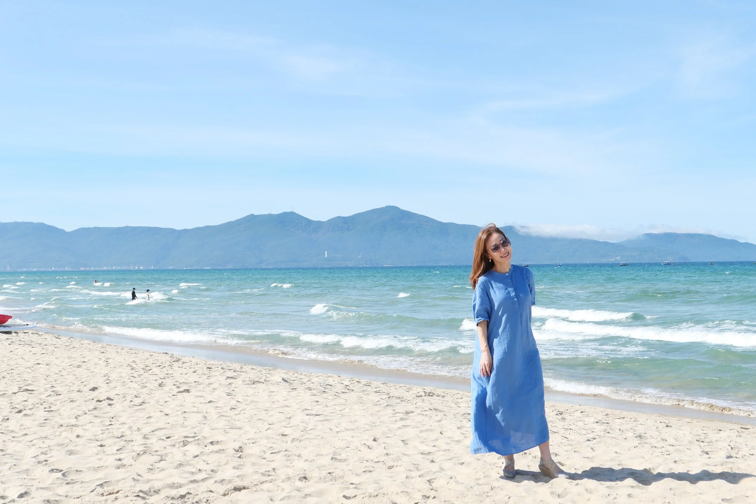 A woman with long hair wearing sunglasses and a blue dress standing on a sandy beach by the ocean, with mountains in the background and a clear sky.
