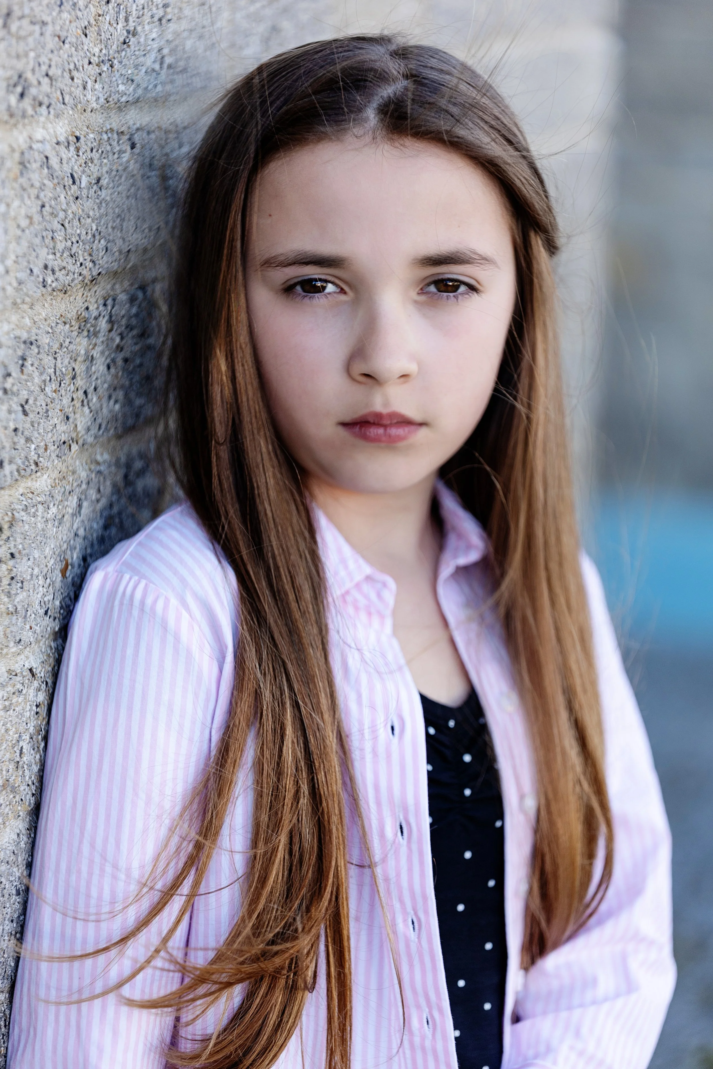 A young girl with long brown hair, brown eyes, wearing a pink and white striped shirt over a black top, leaning against a textured brick wall.