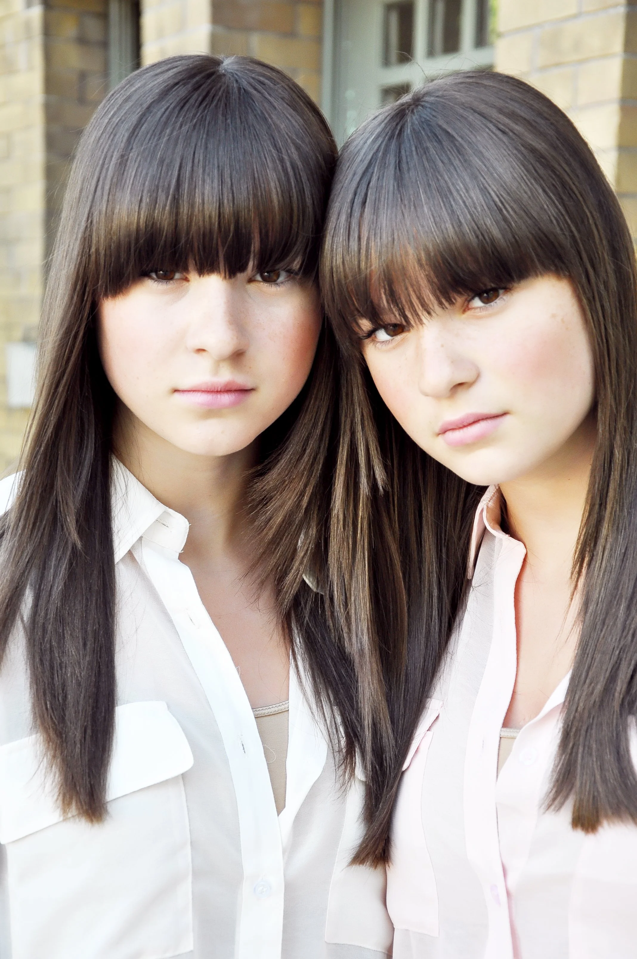 Two young women with long brown hair and blunt bangs, wearing white shirts, appear to be close and posing for a photo outdoors near a brick building.