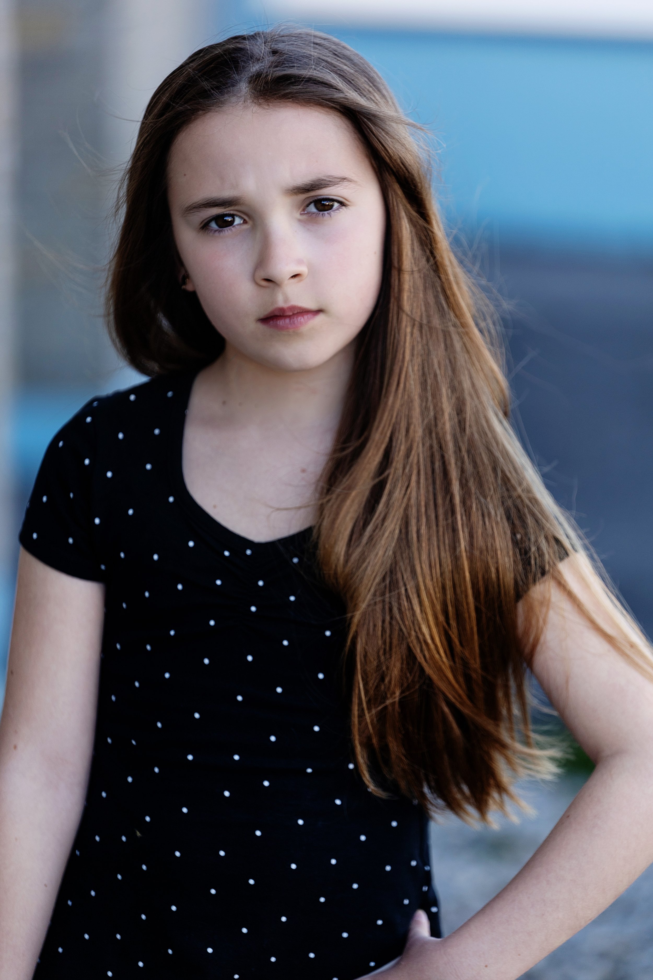 A young girl with long brown hair, wearing a black shirt with white polka dots, looks directly at the camera with a serious expression.