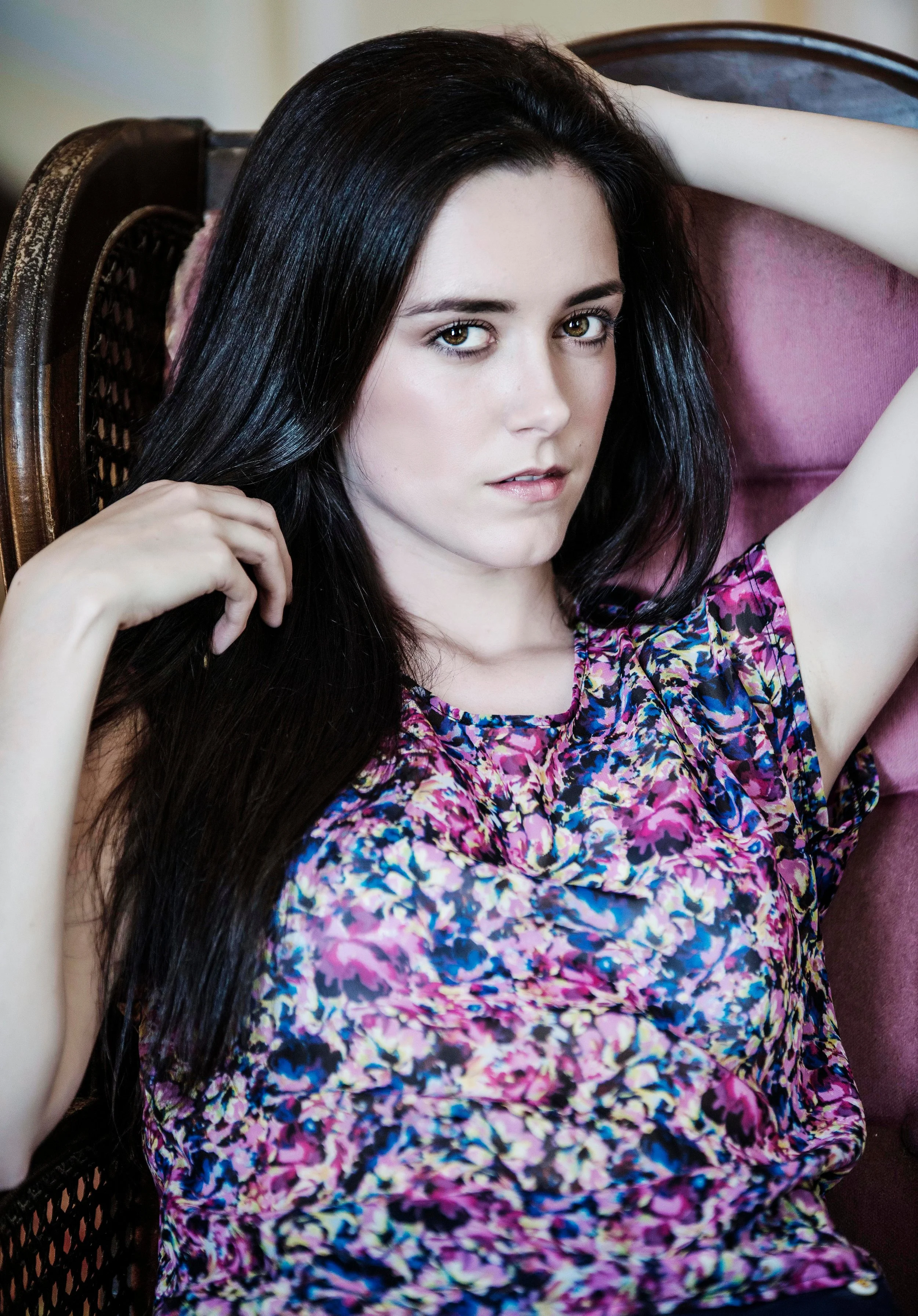 A young woman with long dark hair and fair skin, wearing a colorful, floral-patterned blouse, sitting on a pink cushioned chair with wooden armrests. She is looking at the camera with one arm raised behind her head and the other hand touching her hai