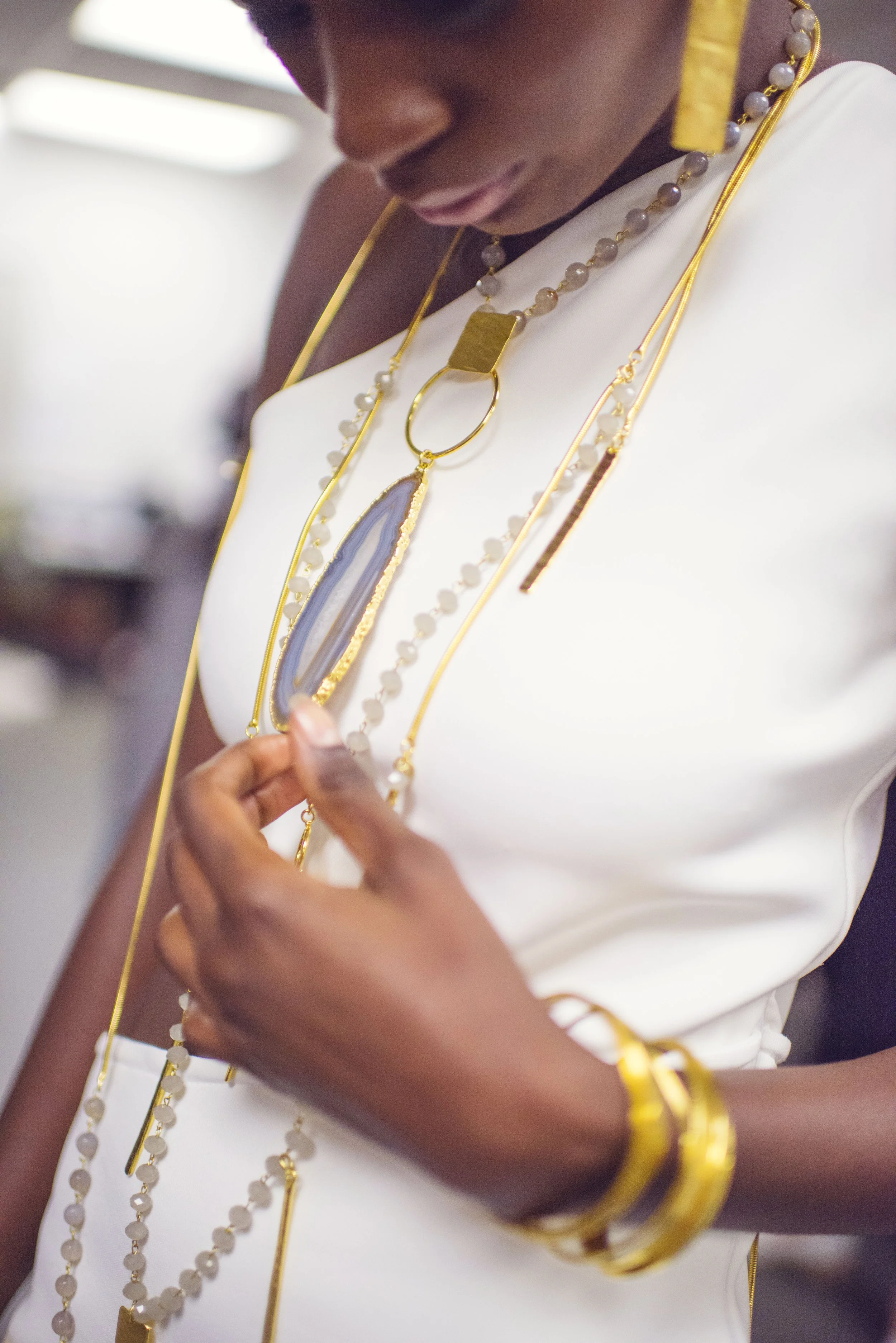 A woman wearing multiple necklaces, including a long one with white beads, a gold chain with a large pendant, and other gold and bead jewelry, dressed in a white top with yellow accents.