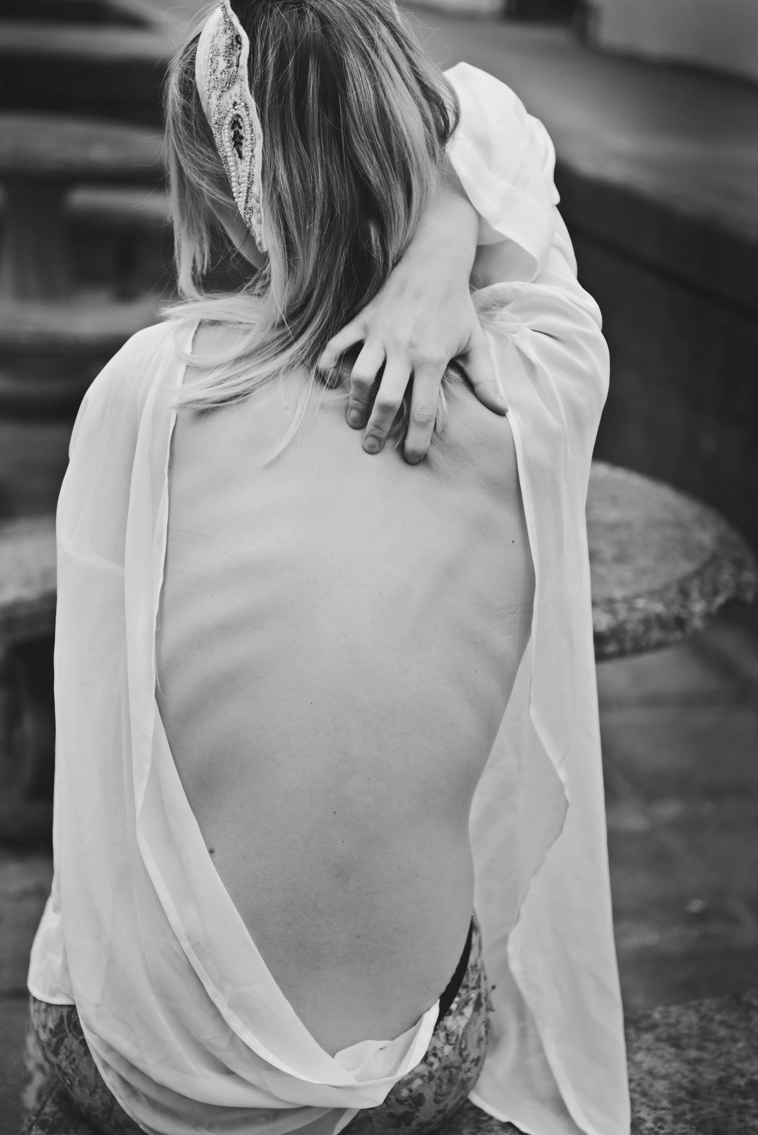 Black and white photo of a person sitting outside. They are wearing a loose, open shirt with one arm resting on their knee and the other hand on their shoulder. The person has long hair and an embellished headband.