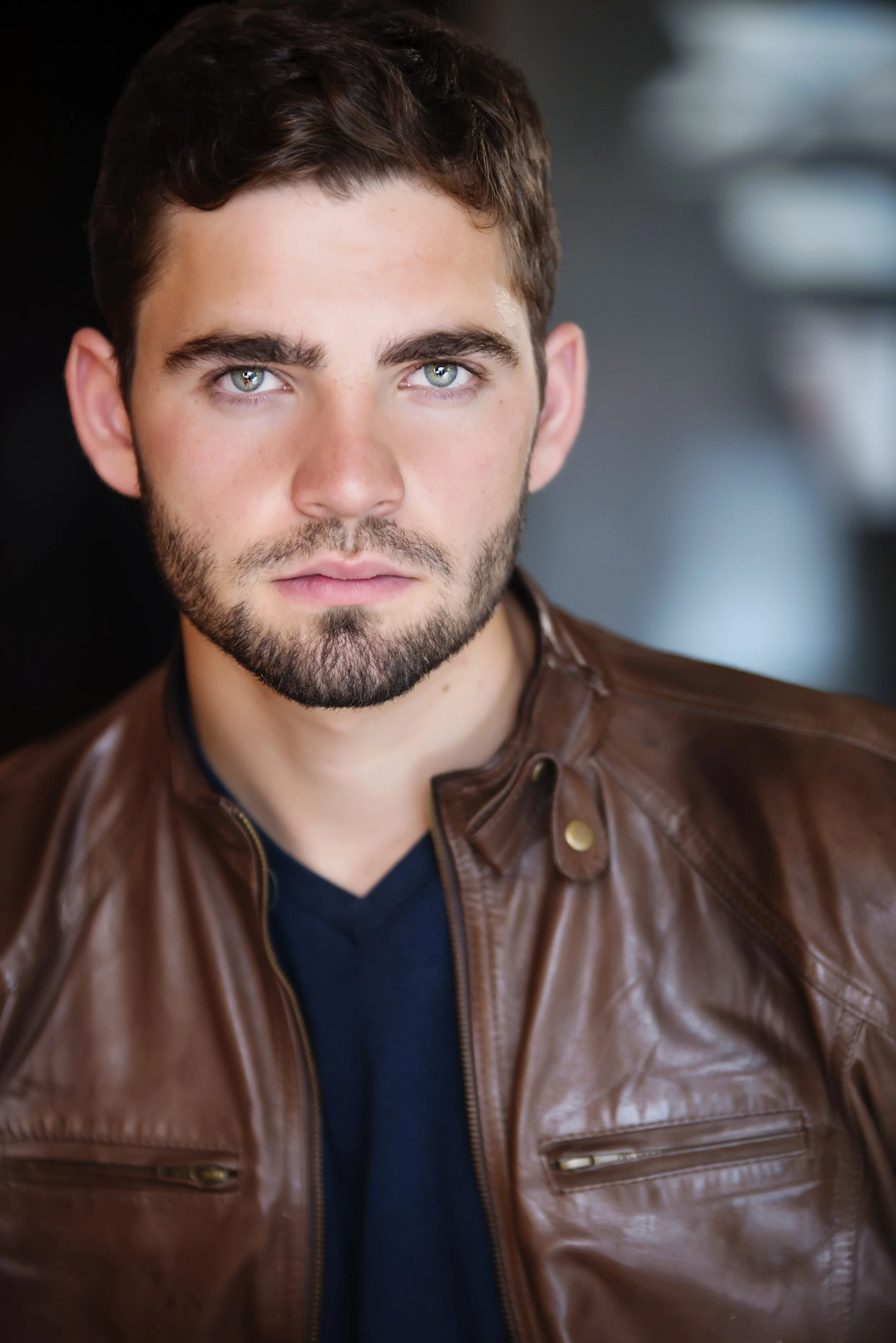 Close-up of a young man with brown hair and blue eyes, wearing a brown leather jacket over a dark shirt, looking directly at the camera.