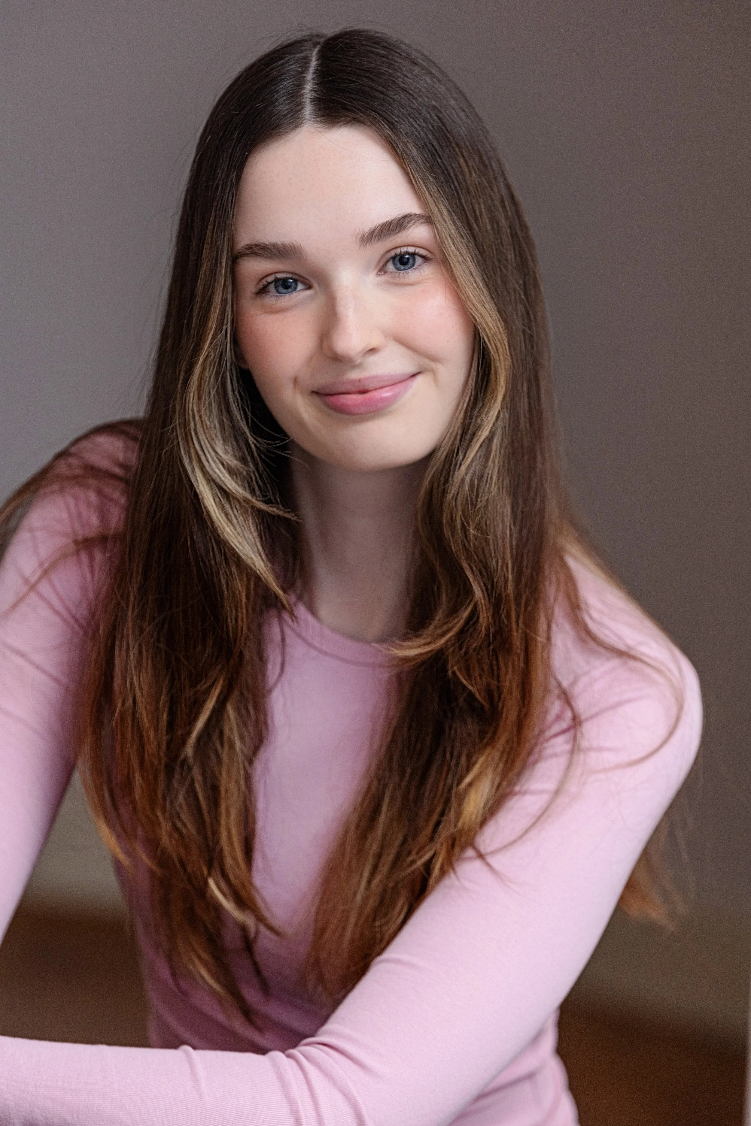 A young woman with long, wavy brown hair and blue eyes, smiling softly, wearing a pink long-sleeve shirt, sitting indoors with a plain gray background.