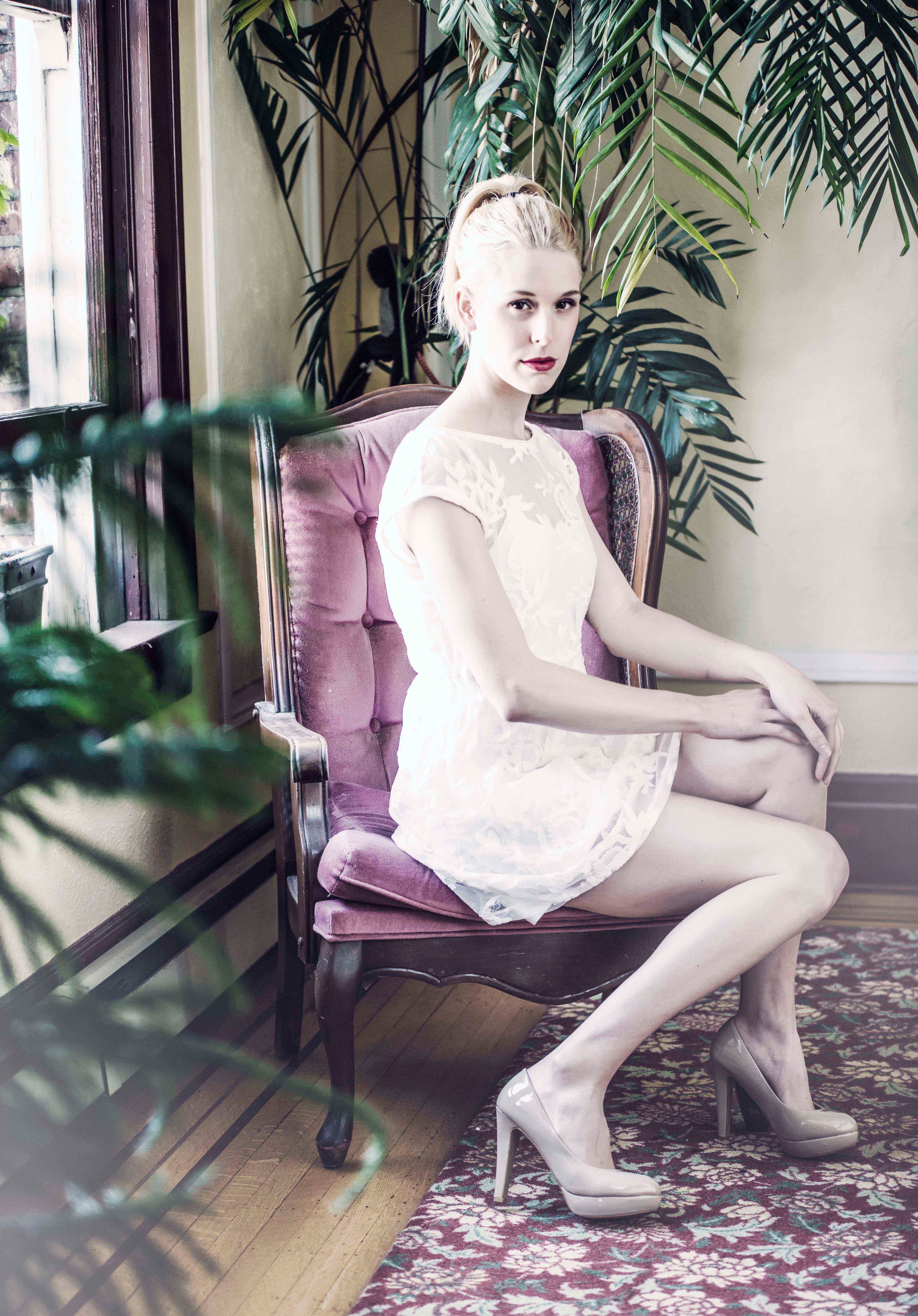 A woman with platinum blonde hair, dressed in a white lace dress, seated on a vintage purple velvet chair with wooden trim. She is wearing high-heeled shoes and sitting with her legs crossed, near a window with a large green plant behind her in a coz