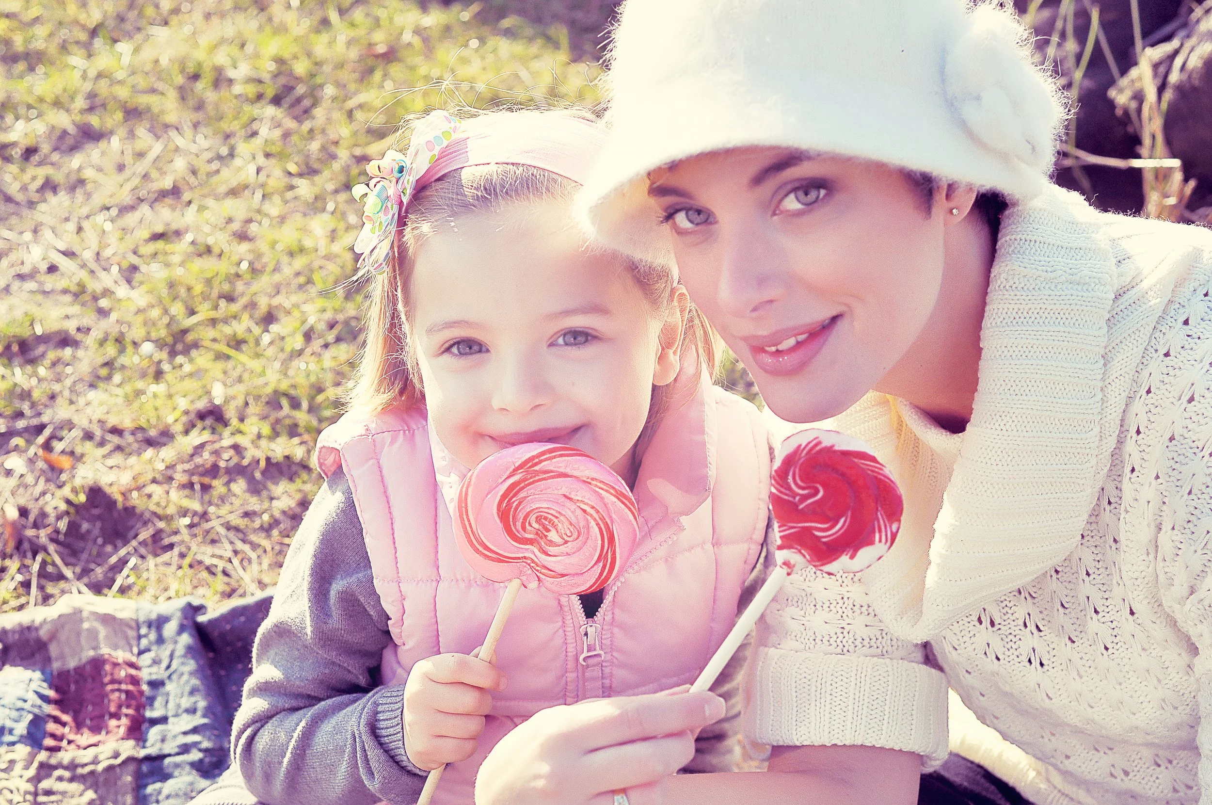 A woman and a young girl are sitting outdoors on a sunny day, smiling, with the girl holding a pink and white swirled lollipop. The woman is wearing a white knit sweater and white hat, and the girl is wearing a pink jacket and a colorful polka-dot he