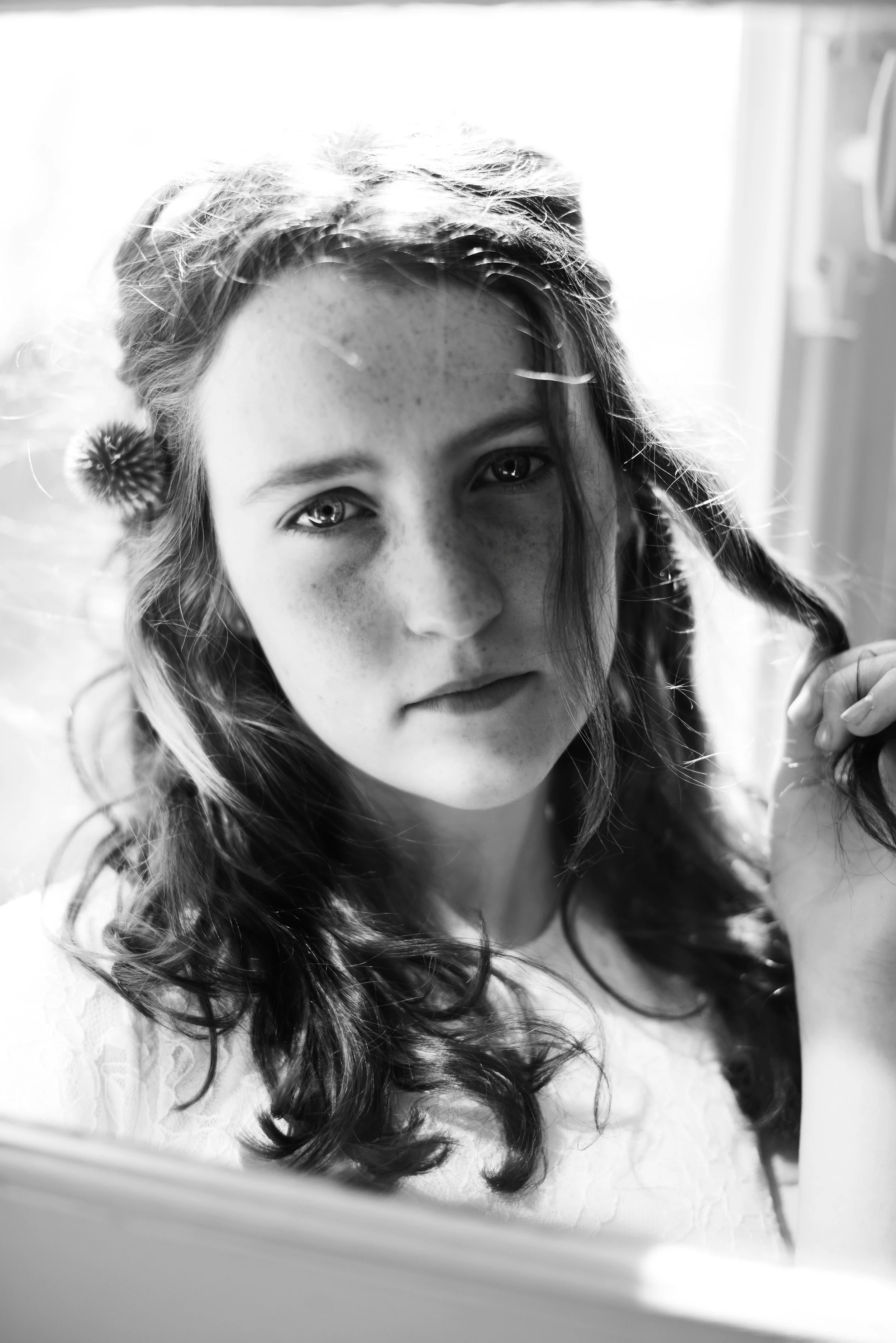 Black and white photo of a young girl with long curly hair, freckles, and light eyes, seen through a mirror with a reflection of her face and part of her hand.