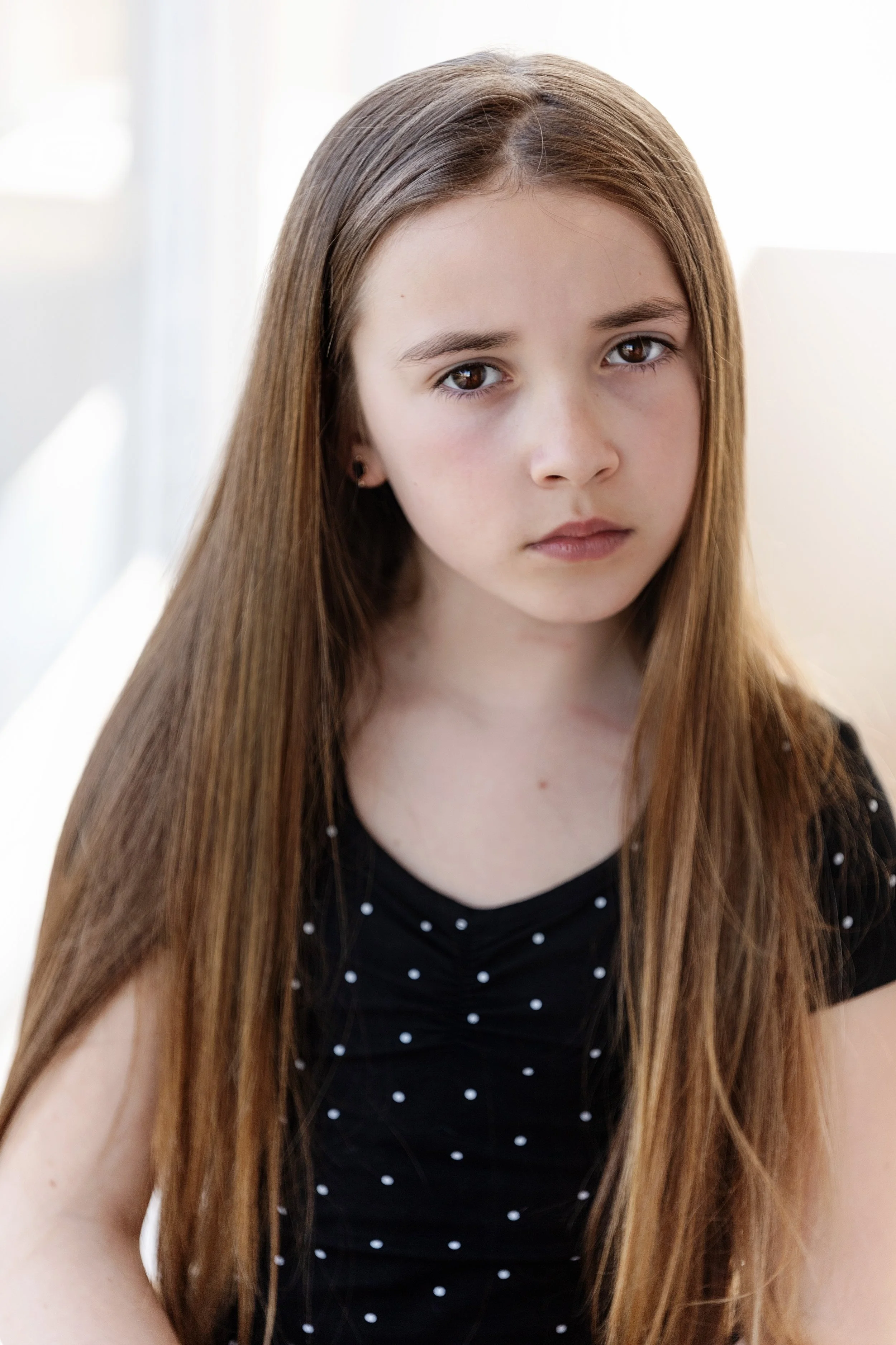 A young girl with long, straight brown hair and brown eyes, wearing a black shirt with white polka dots, looking directly at the camera.