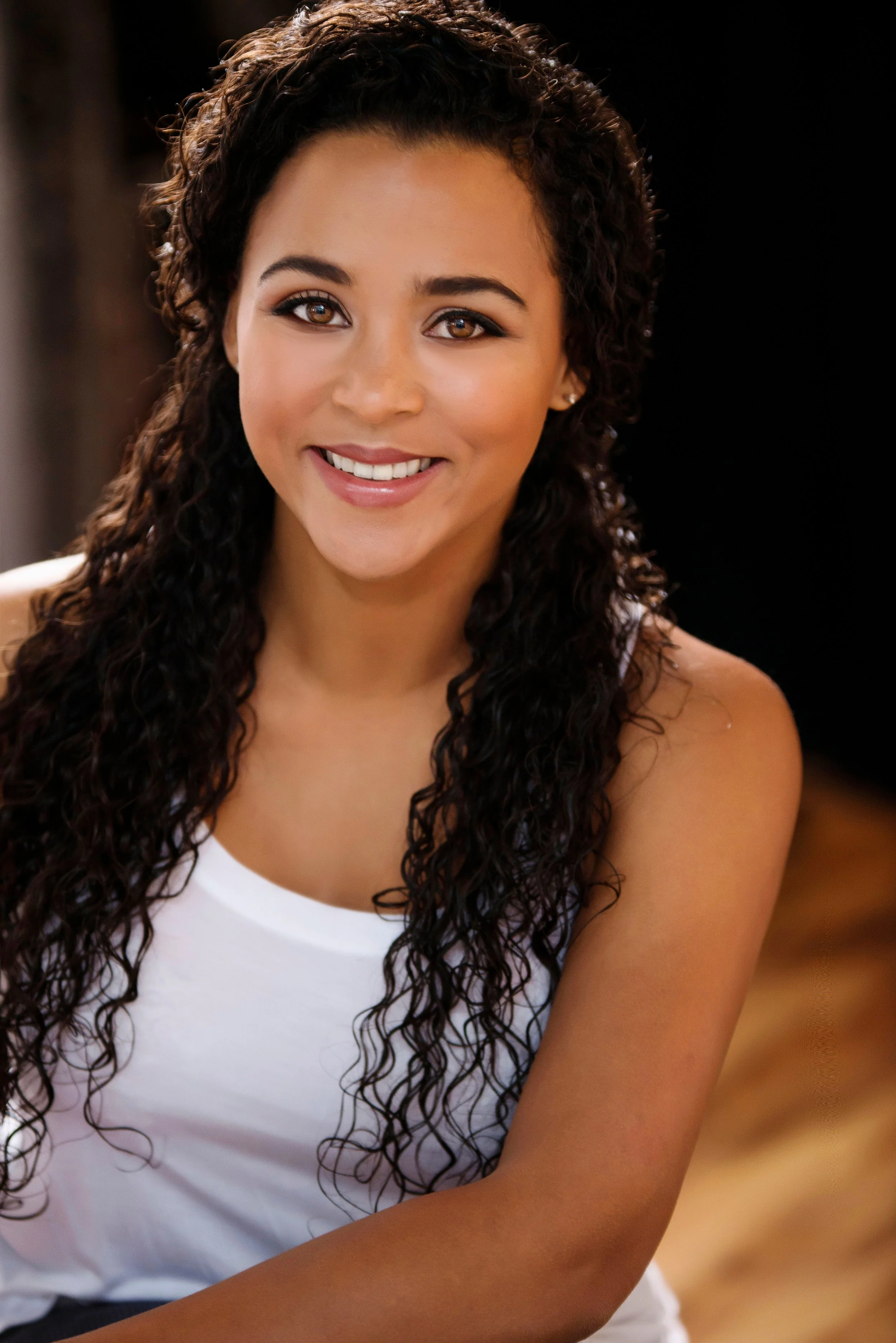 Close-up portrait of a young woman with curly dark brown hair, brown eyes, and a bright smile, wearing a white tank top, against a dark background.