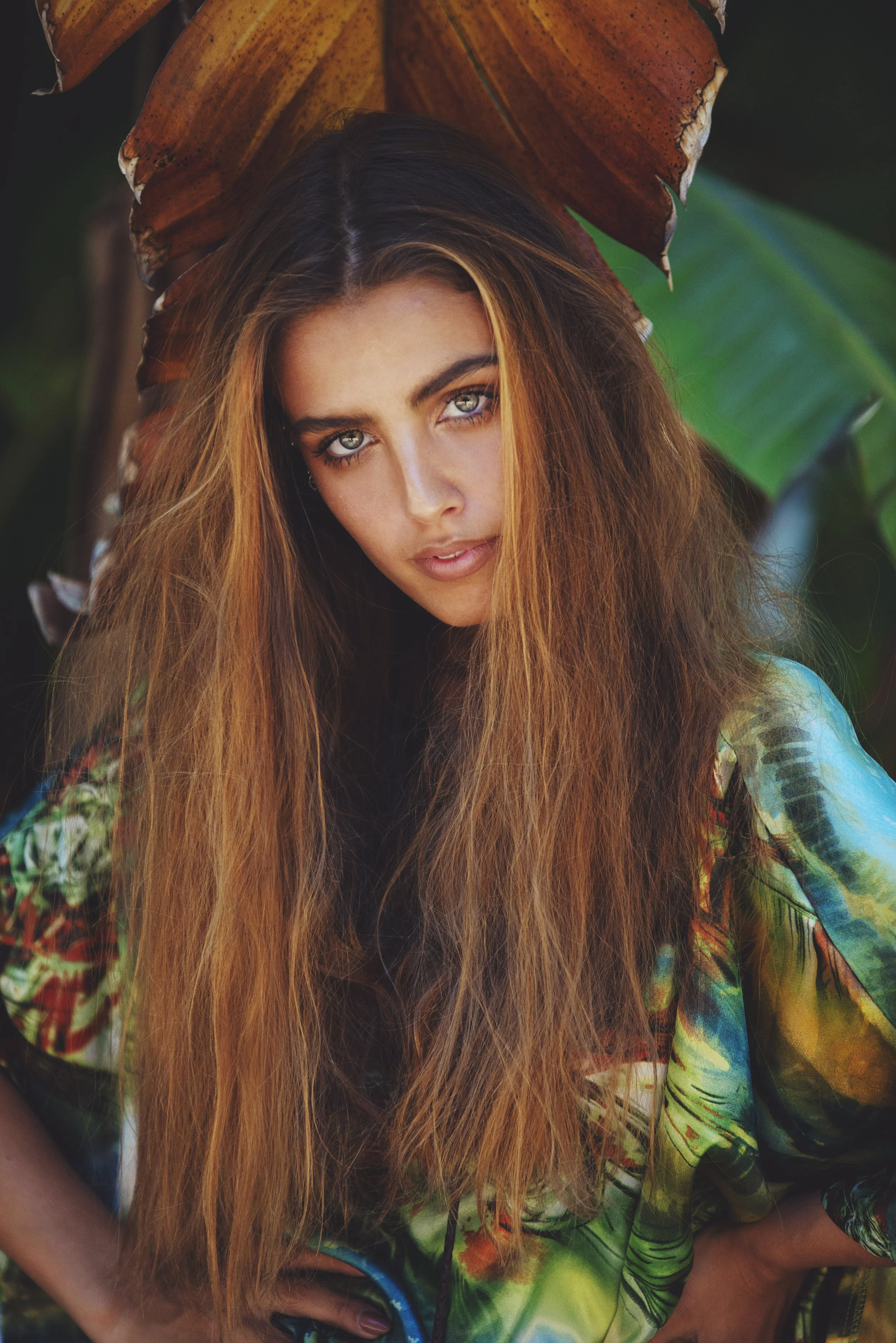 Close-up of a young woman with long, wavy hair and striking blue-green eyes, wearing a colorful, patterned top, standing outdoors with large tropical leaves in the background.