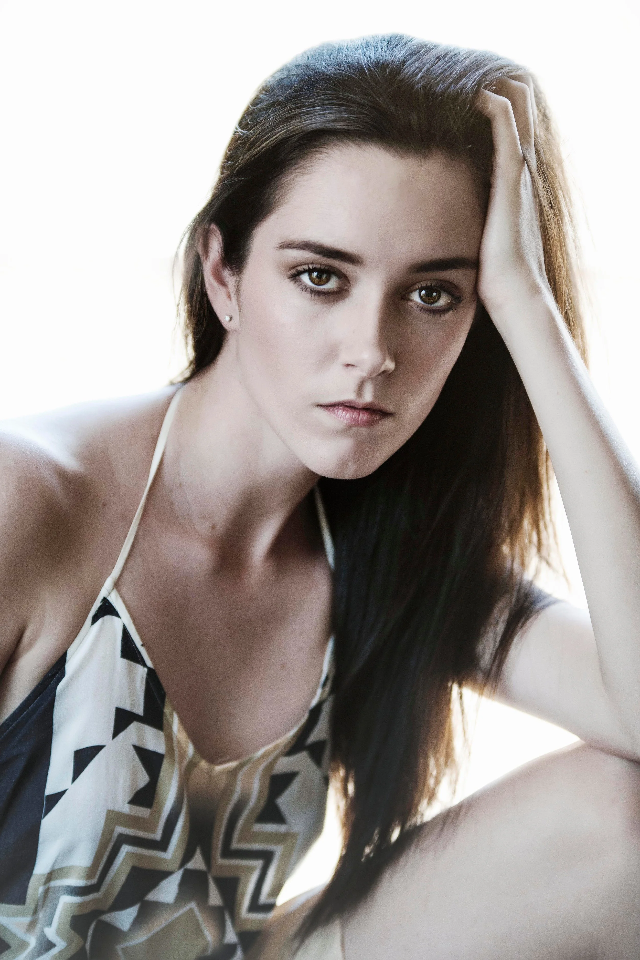 A young woman with long brown hair and hazel eyes, resting her head on her hand, wearing a black and white patterned dress, posing indoors with natural light.