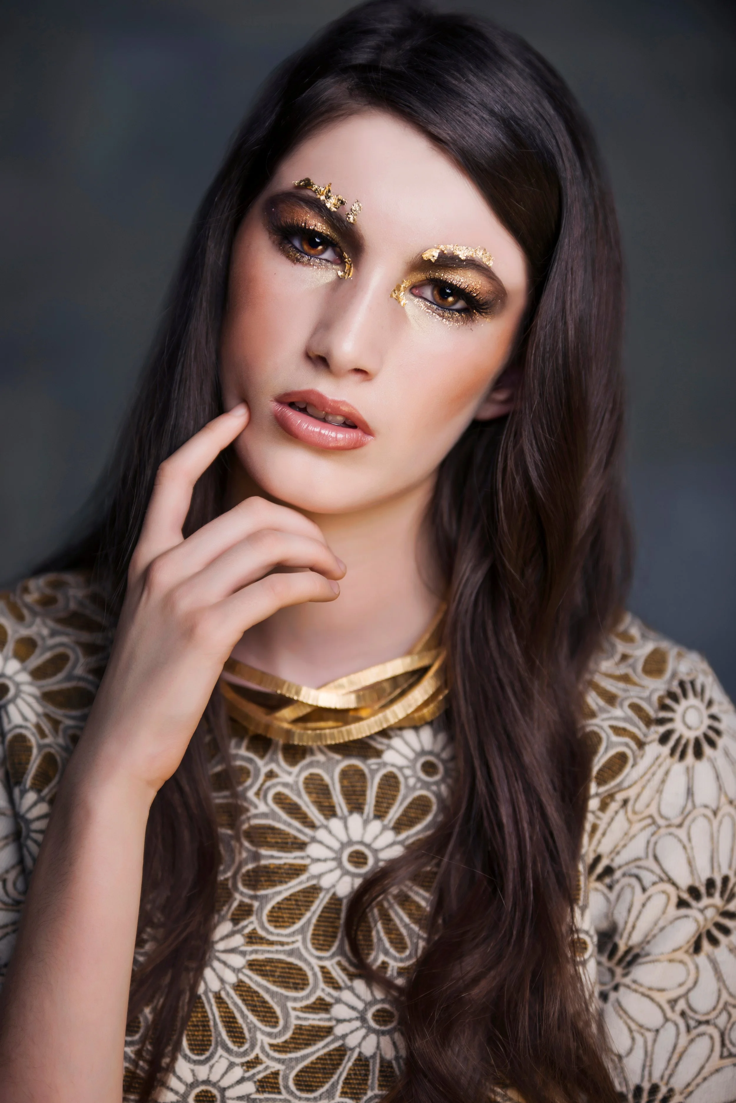 A woman with long brown hair wearing gold and glitter makeup, gold embellishments near her eyes, a patterned dress with floral designs, and a layered gold necklace.
