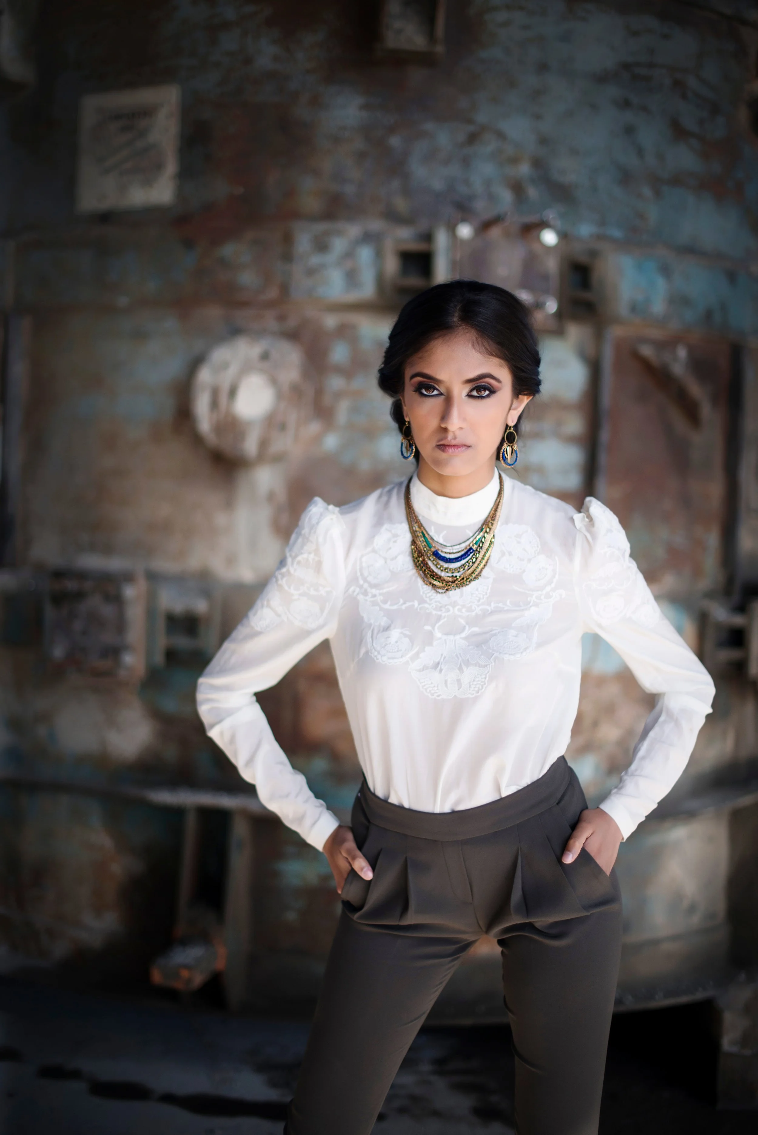 A woman standing with her hands on her hips in front of a rustic brick wall, wearing a white embroidered blouse, dark trousers, and layered necklaces, with earrings, and dark eye makeup.