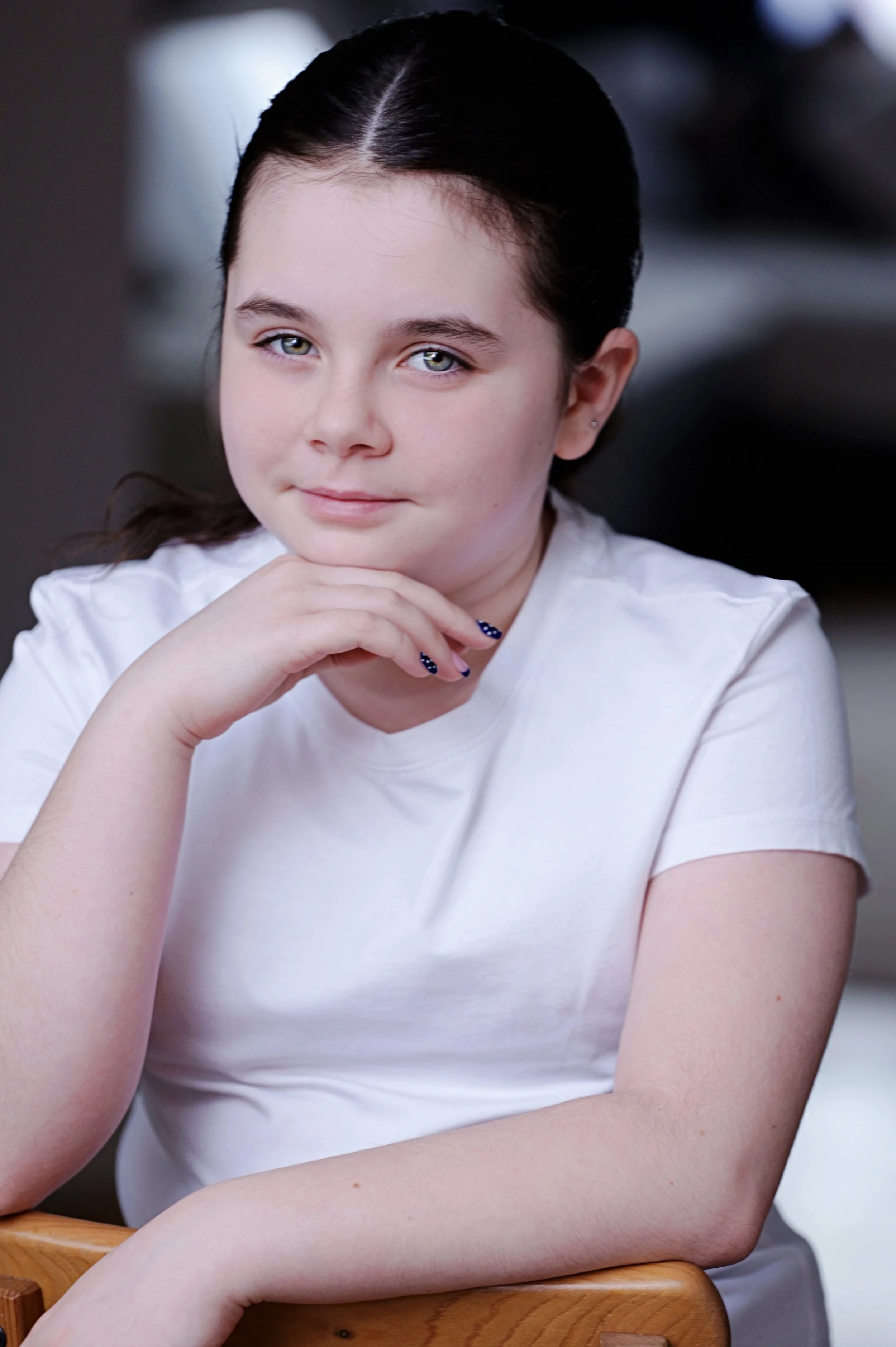 Close-up of a young girl with black hair, green eyes, wearing a white shirt, resting her chin on her hand, with a neutral background.