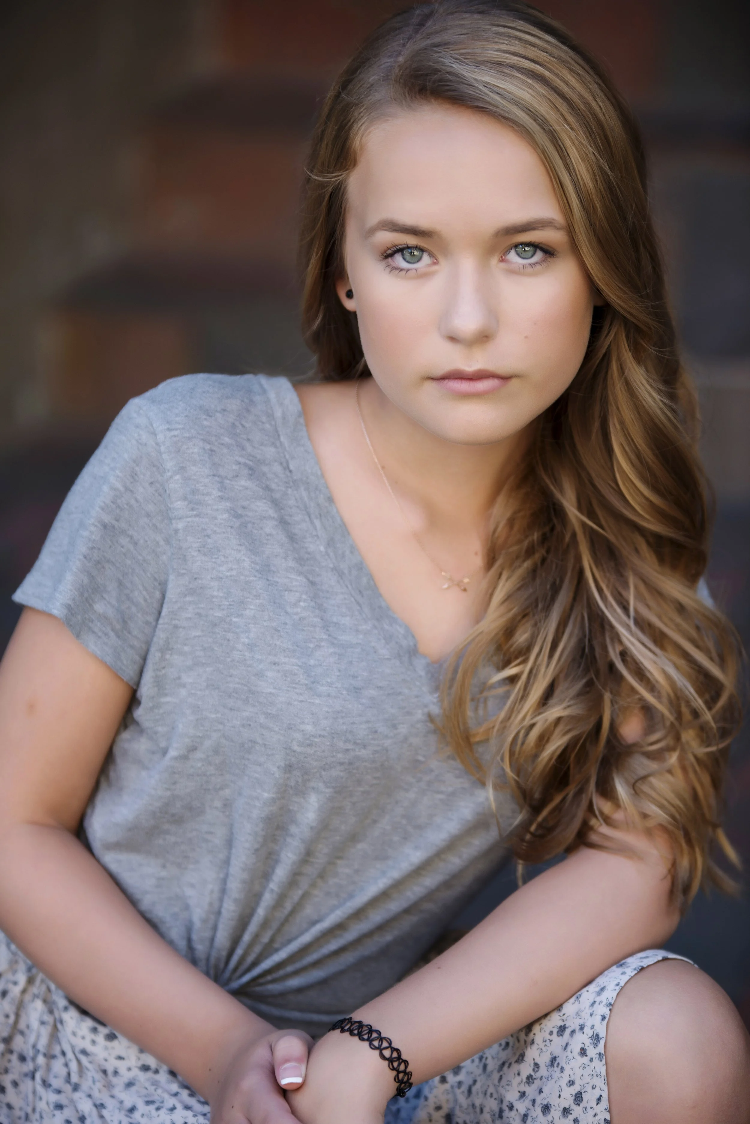 Close-up portrait of a young girl with long, wavy brown hair, blue eyes, wearing a light gray t-shirt, sitting outdoors with arms resting on her knees, and a neutral expression.