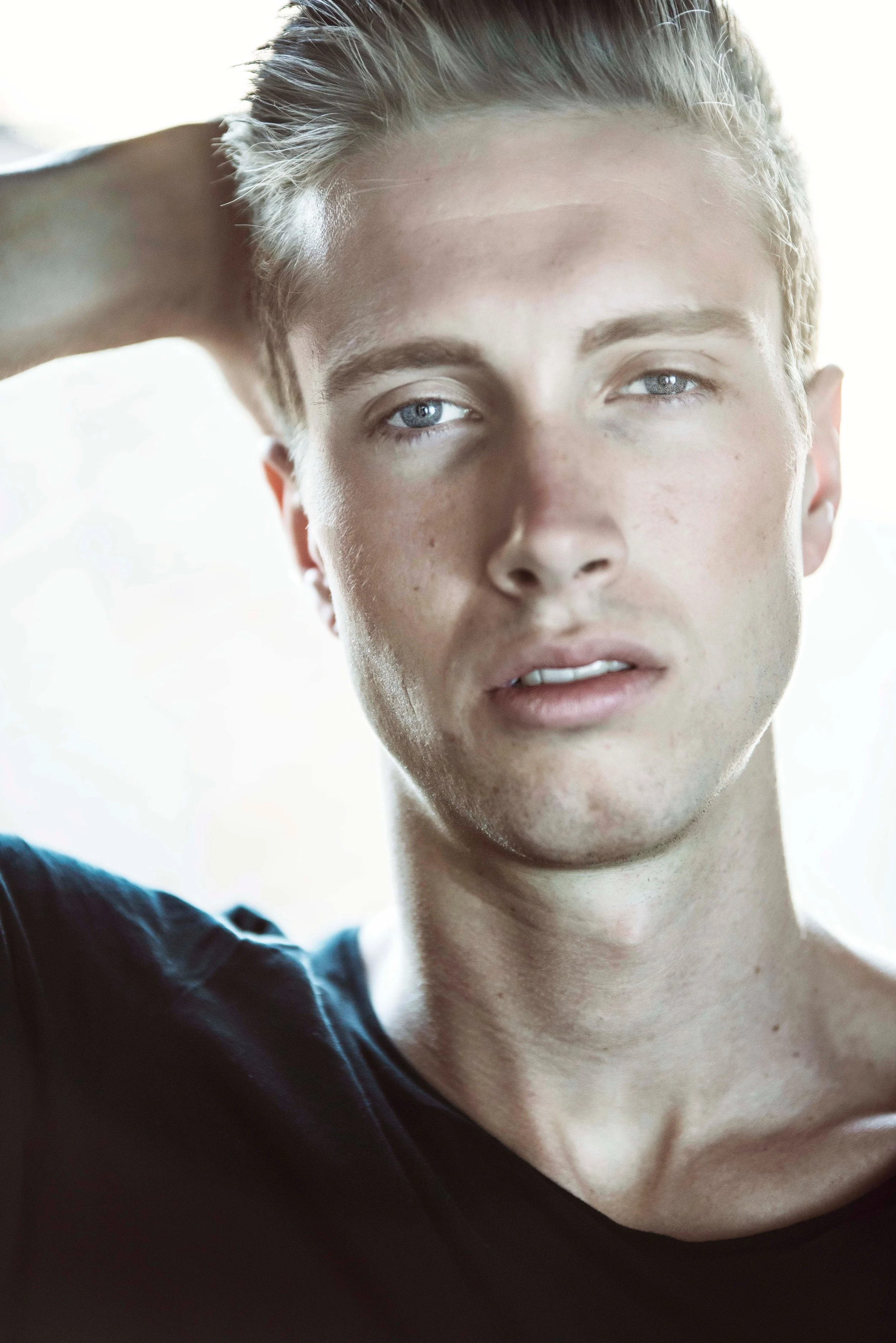 Close-up of a young man with short, light hair and blue eyes in natural light, wearing a dark shirt, with his hand resting behind his head.