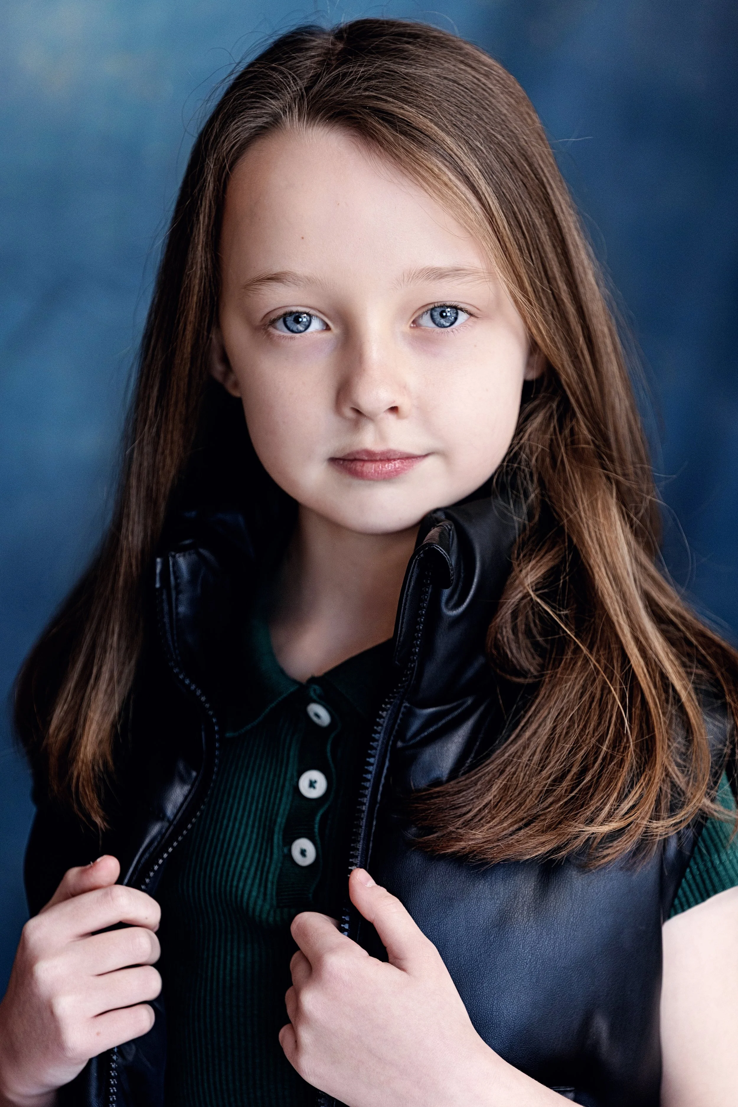 A young girl with long, light brown hair and blue eyes, wearing a dark green shirt and a black jacket, looking directly at the camera with a neutral expression.