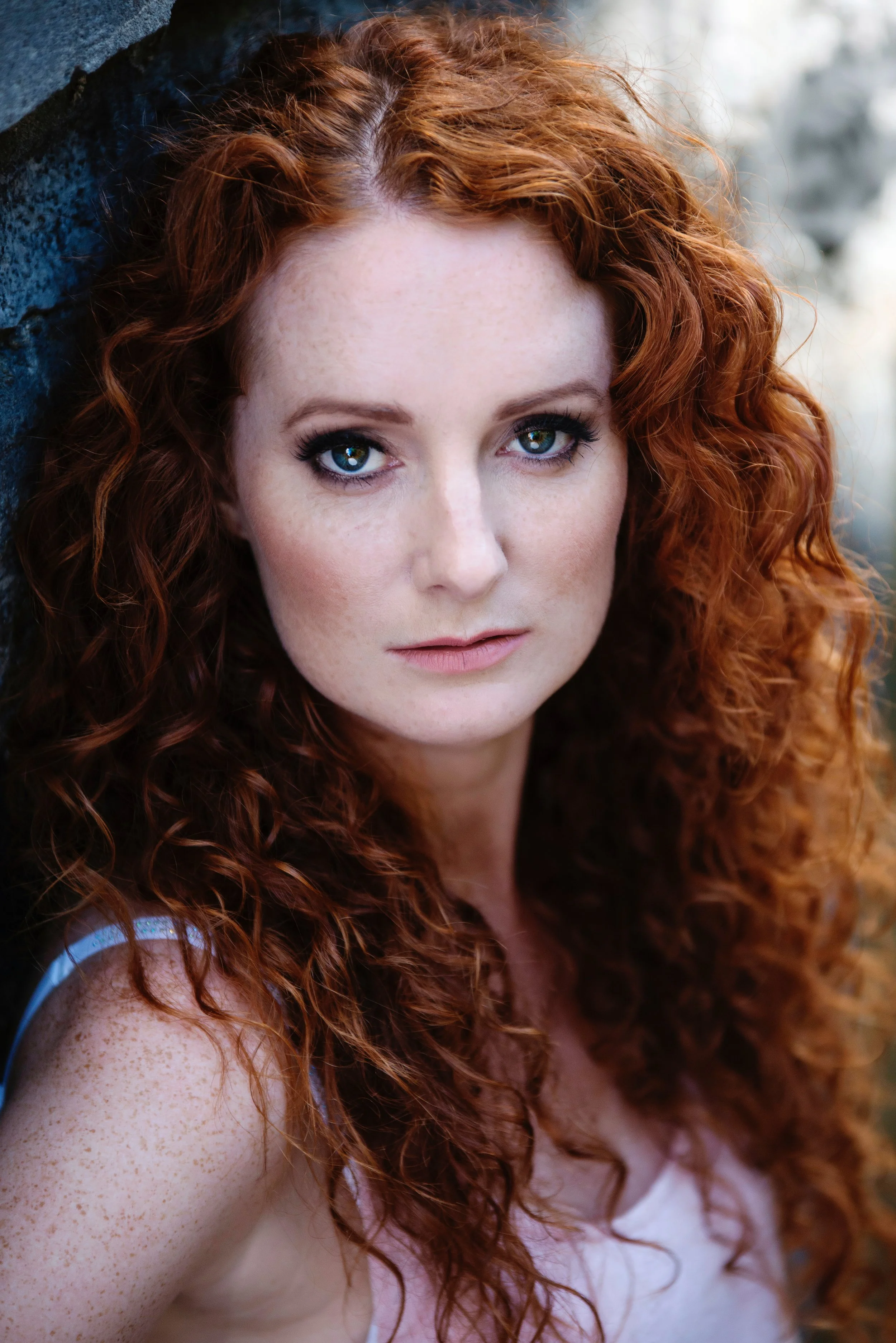 Close-up of a woman with curly red hair and fair skin with freckles, looking directly at the camera, outdoors near a stone wall.