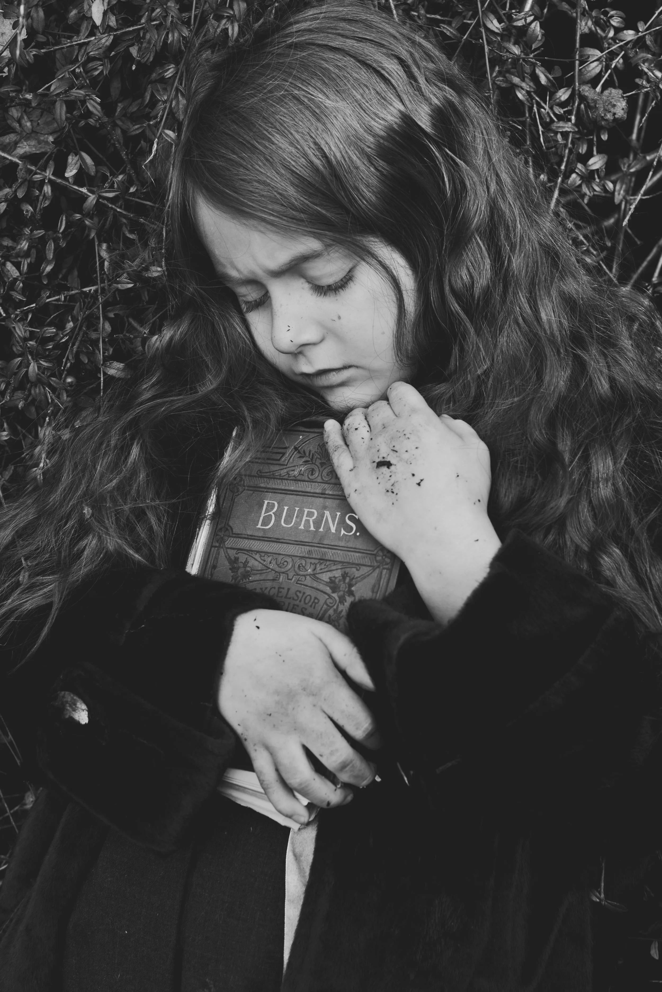 A young woman with long, curly hair lying on the ground, with her eyes closed, surrounded by foliage, holding a book titled 'Burns' and a small stuffed animal.