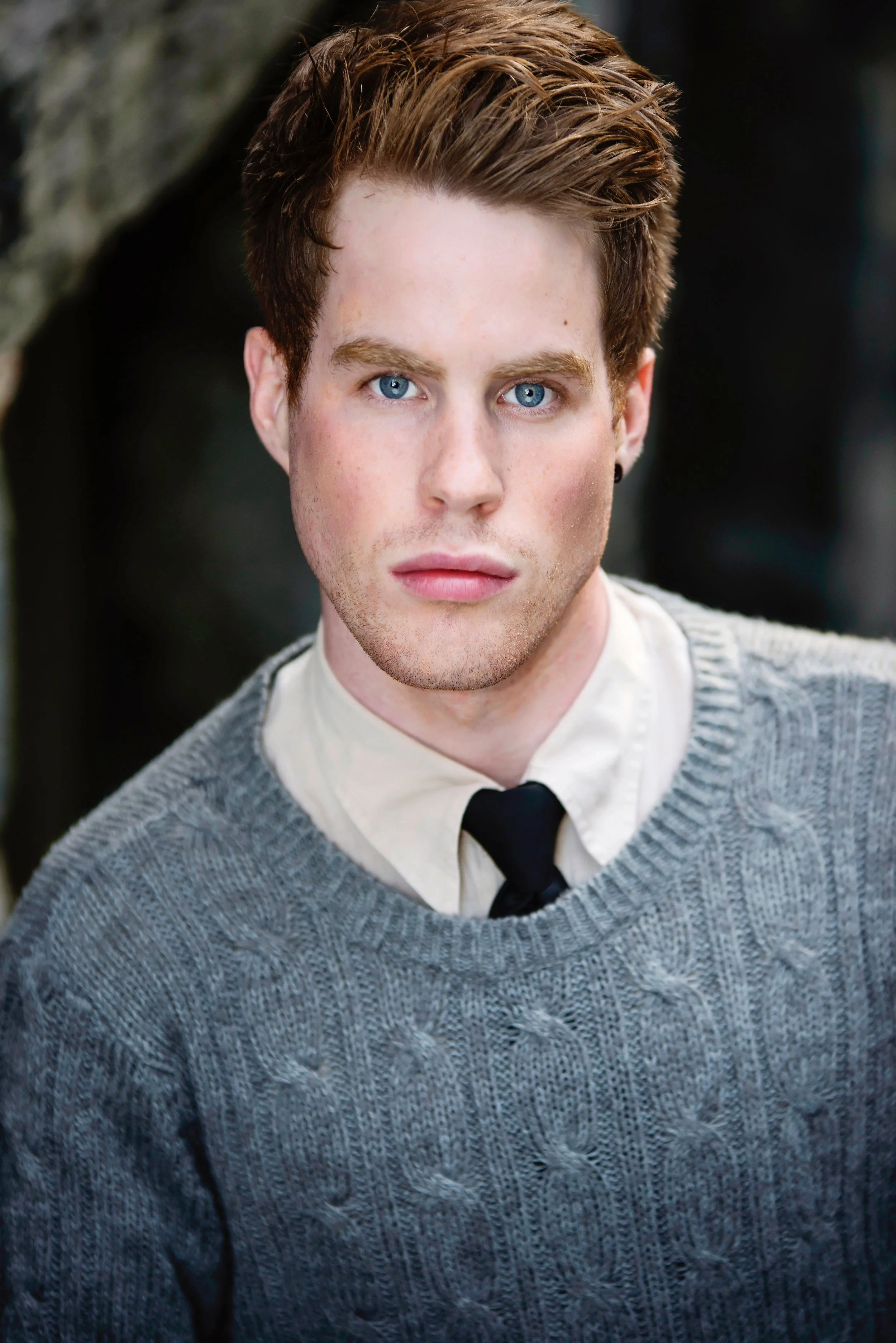 Close-up portrait of a young man with blue eyes, brown hair, fair skin, wearing a white shirt, black tie, and gray knitted sweater.
