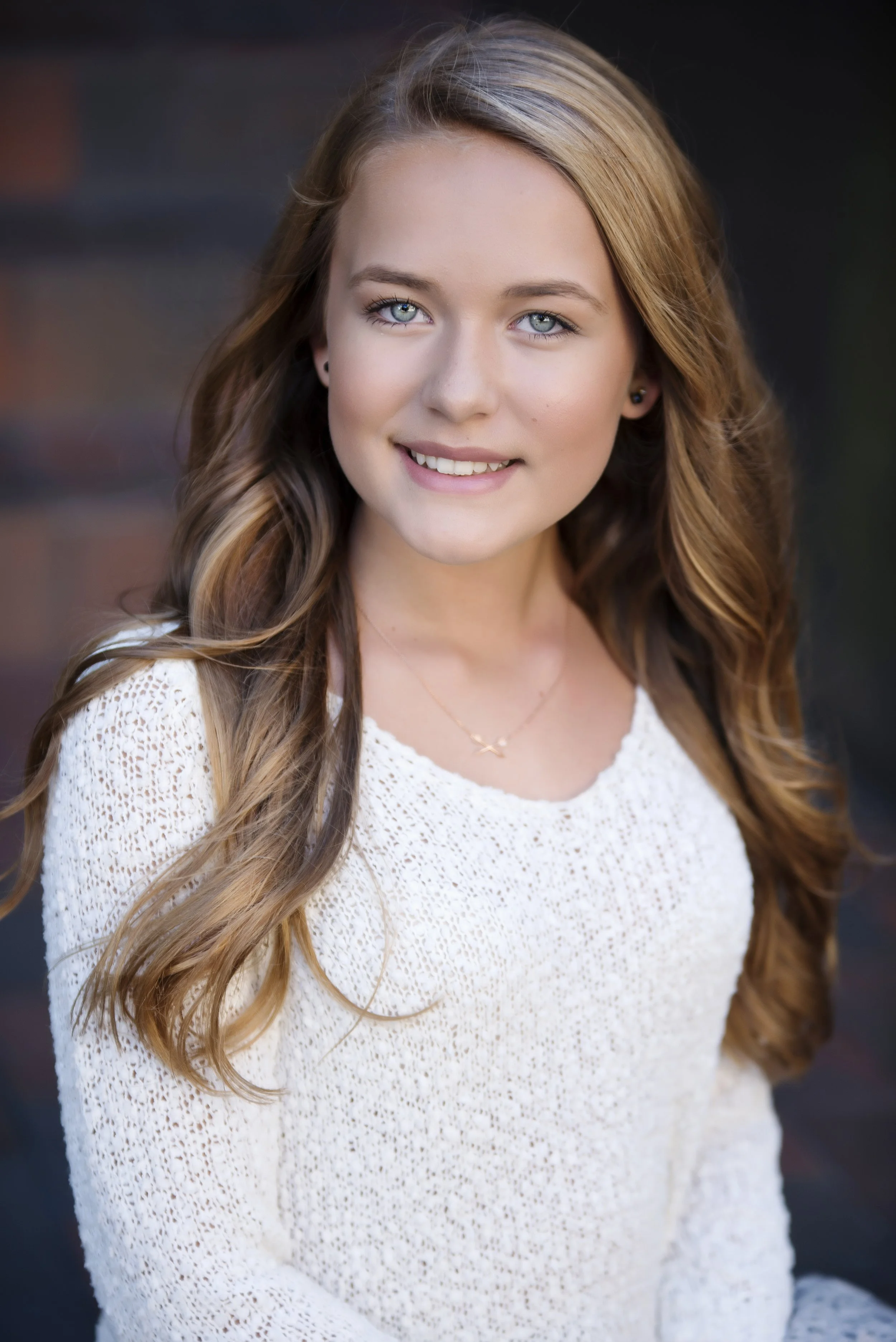 A young woman with long, wavy brown hair and blue eyes is smiling at the camera. She is wearing a white, crocheted top and small earrings, with a delicate gold necklace. The background is blurred and dark.