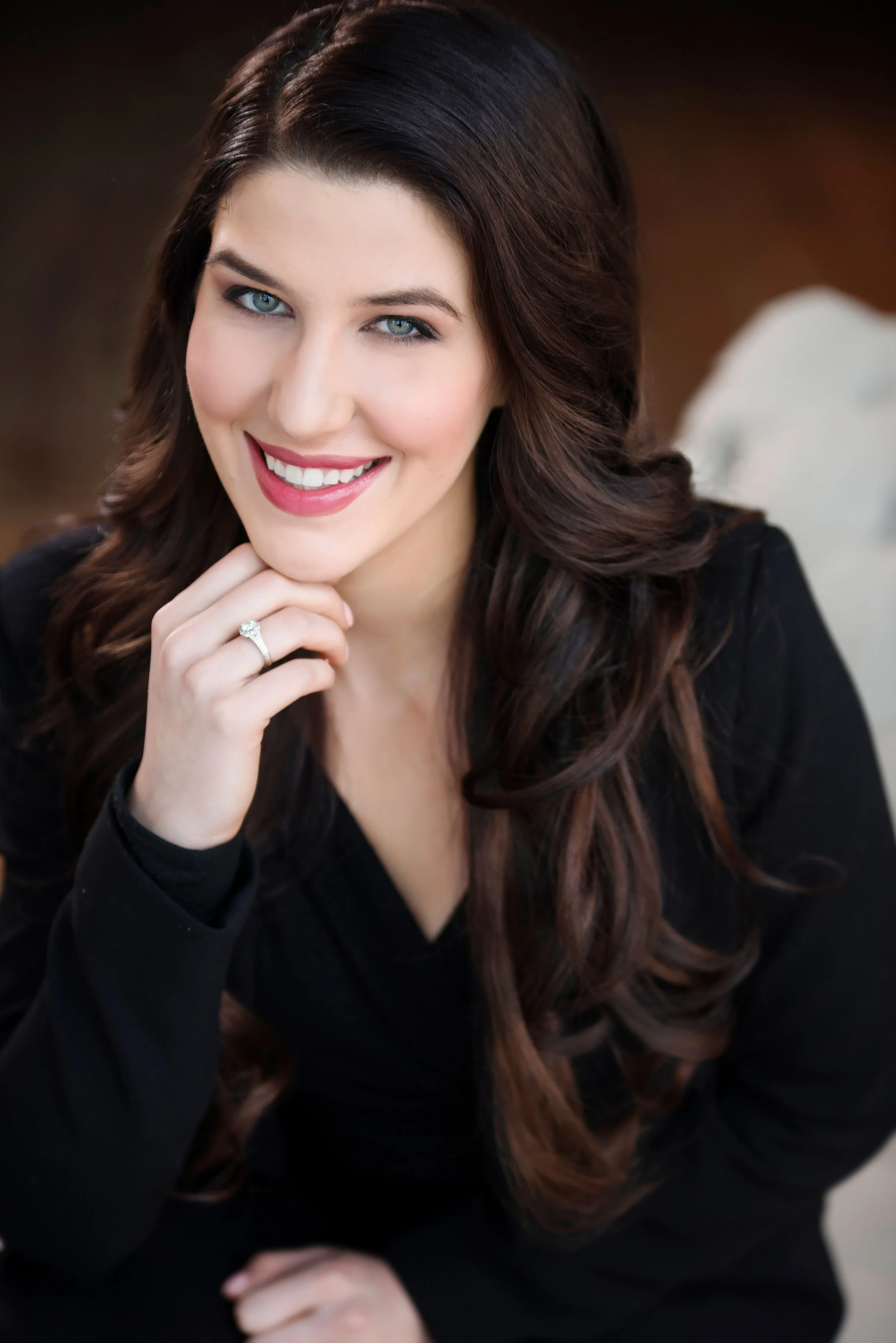 A woman with long wavy brown hair, blue eyes, and a bright smile, wearing a black top and a diamond engagement ring on her left hand, resting her chin on her left hand.